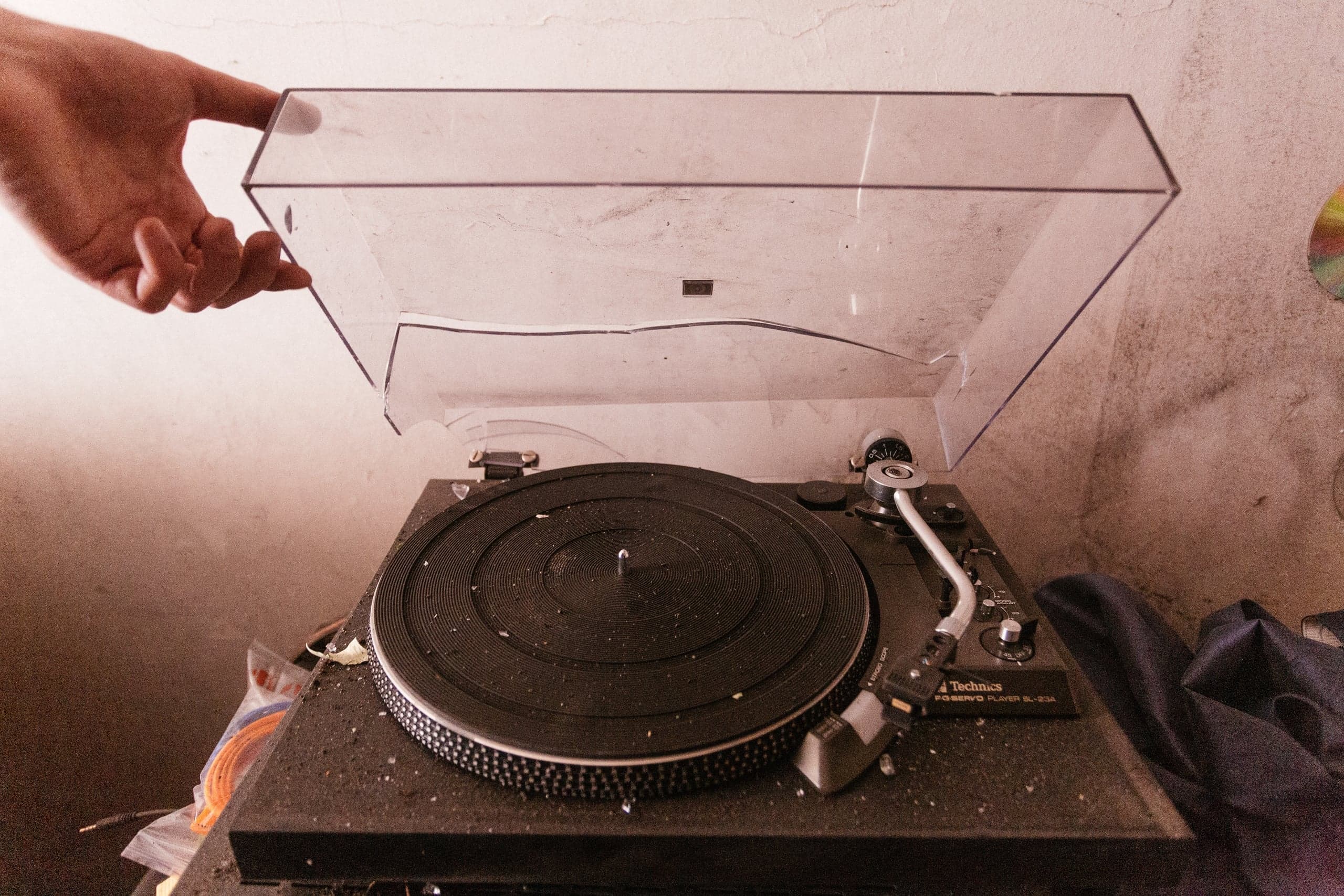Рука открывает треснувшую прозрачную крышку сильно запыленного винилового проигрывателя Technics SL-23A.