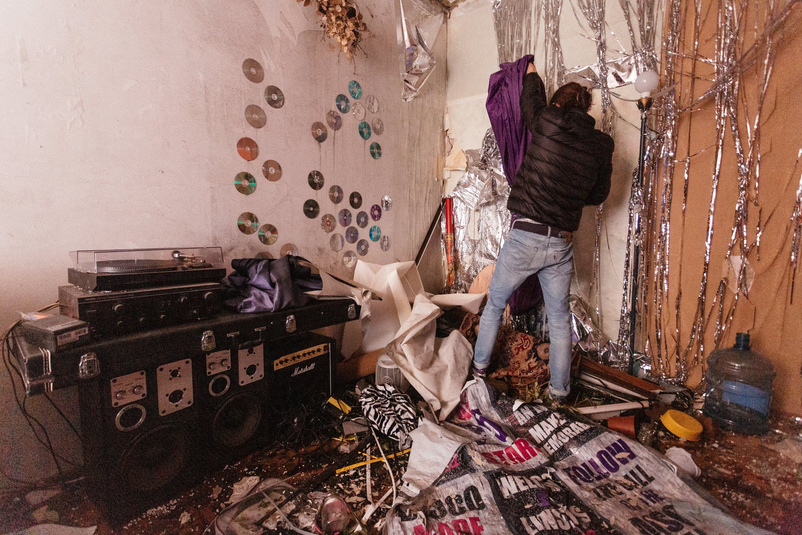 Крупный план: человек в черной куртке и джинсах стоит в разрушенной, заваленной мусором комнате, увешанной серебристой мишурой. Слева на стене закреплены компакт-диски, под ними — старая стереосистема с проигрывателем.