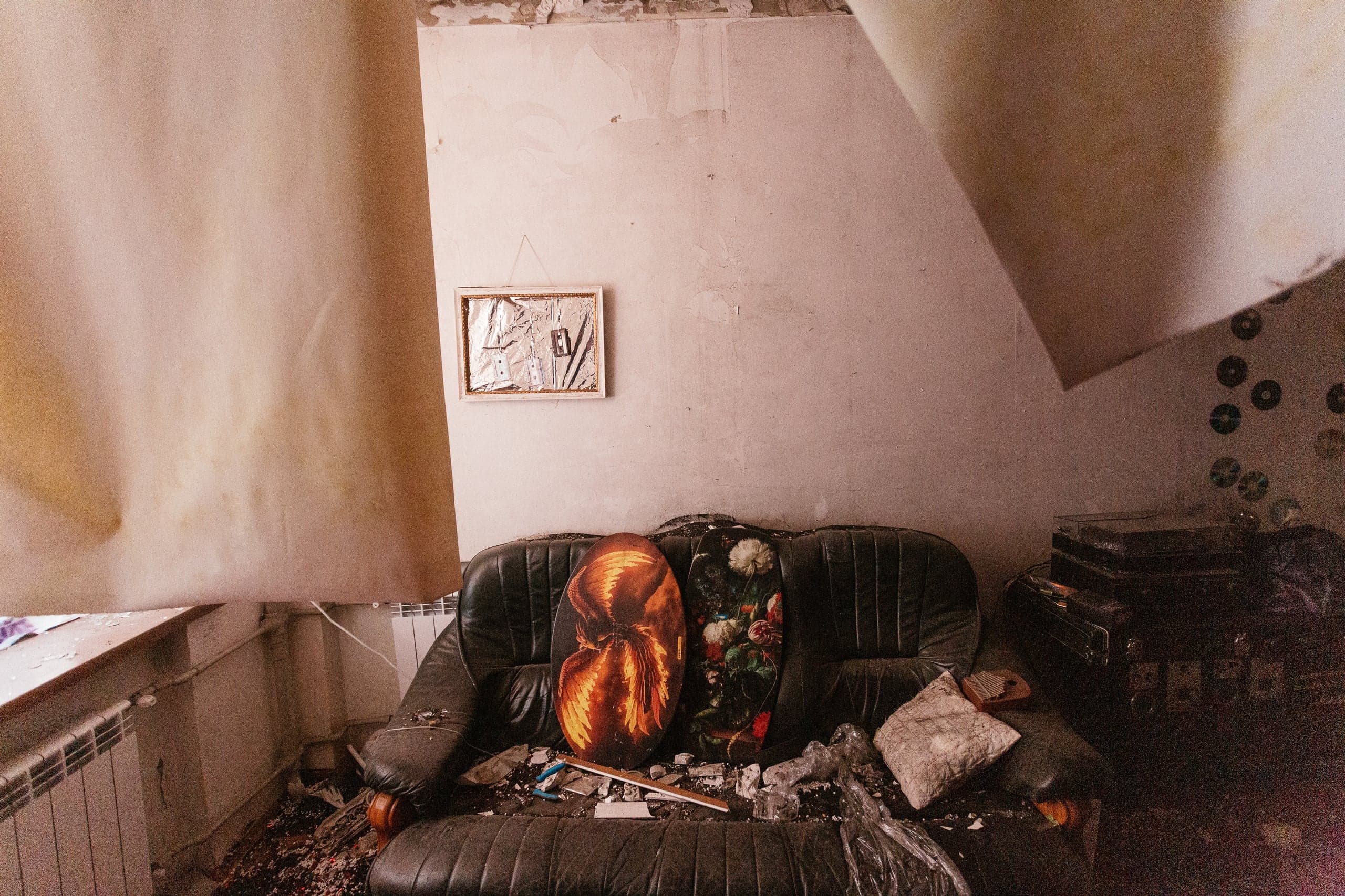 Разрушенная комната: черный диван завален мусором и обломками, а с потолка свисает большой кусок материала. Над диваном висит небольшая картина.