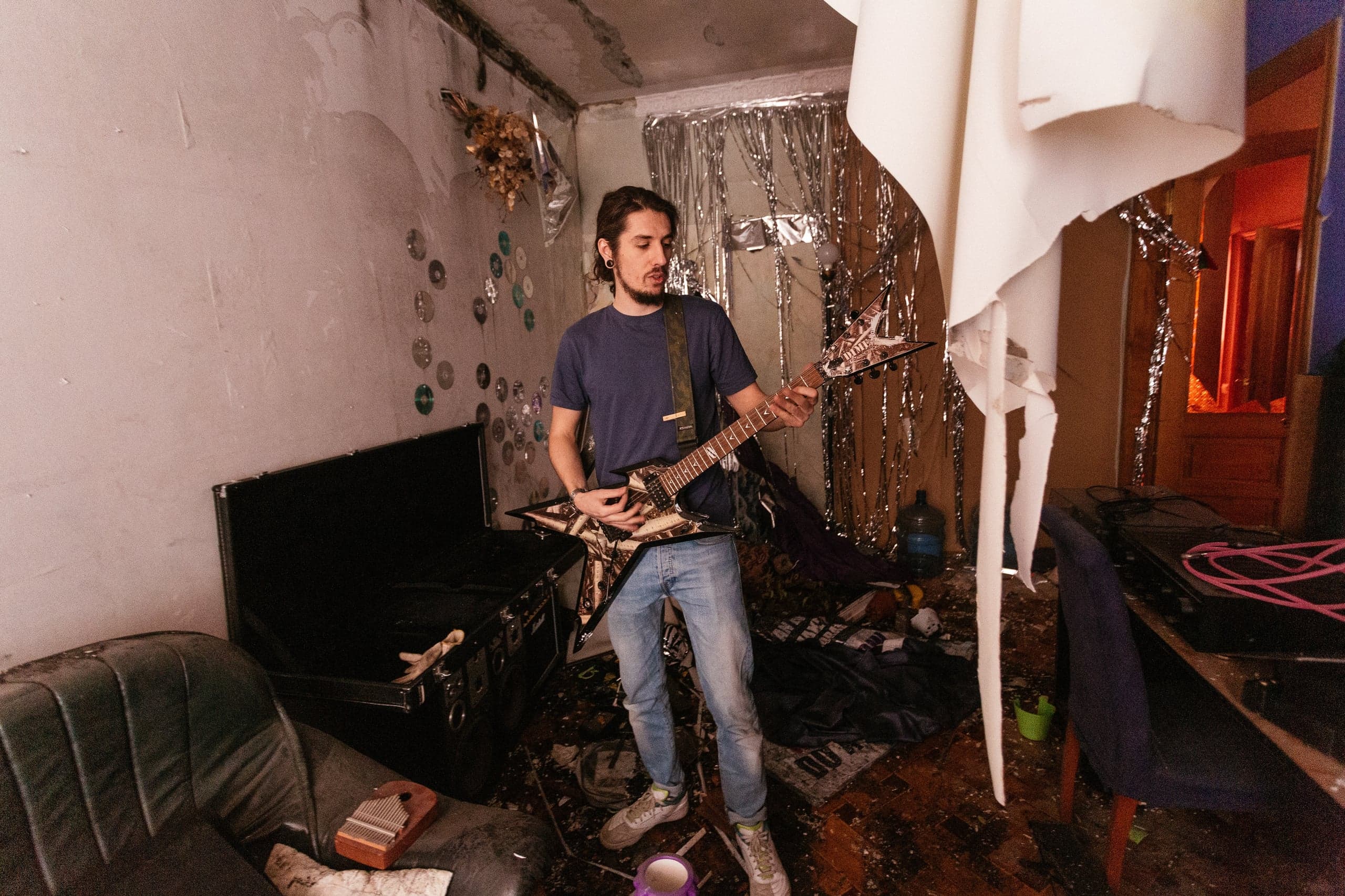 Молодой музыкант играет на электрогитаре Dimebag Dean в захламленной комнате, стены которой украшены приклеенными компакт-дисками и серебряной мишурой. Справа свисает кусок светлого материала.