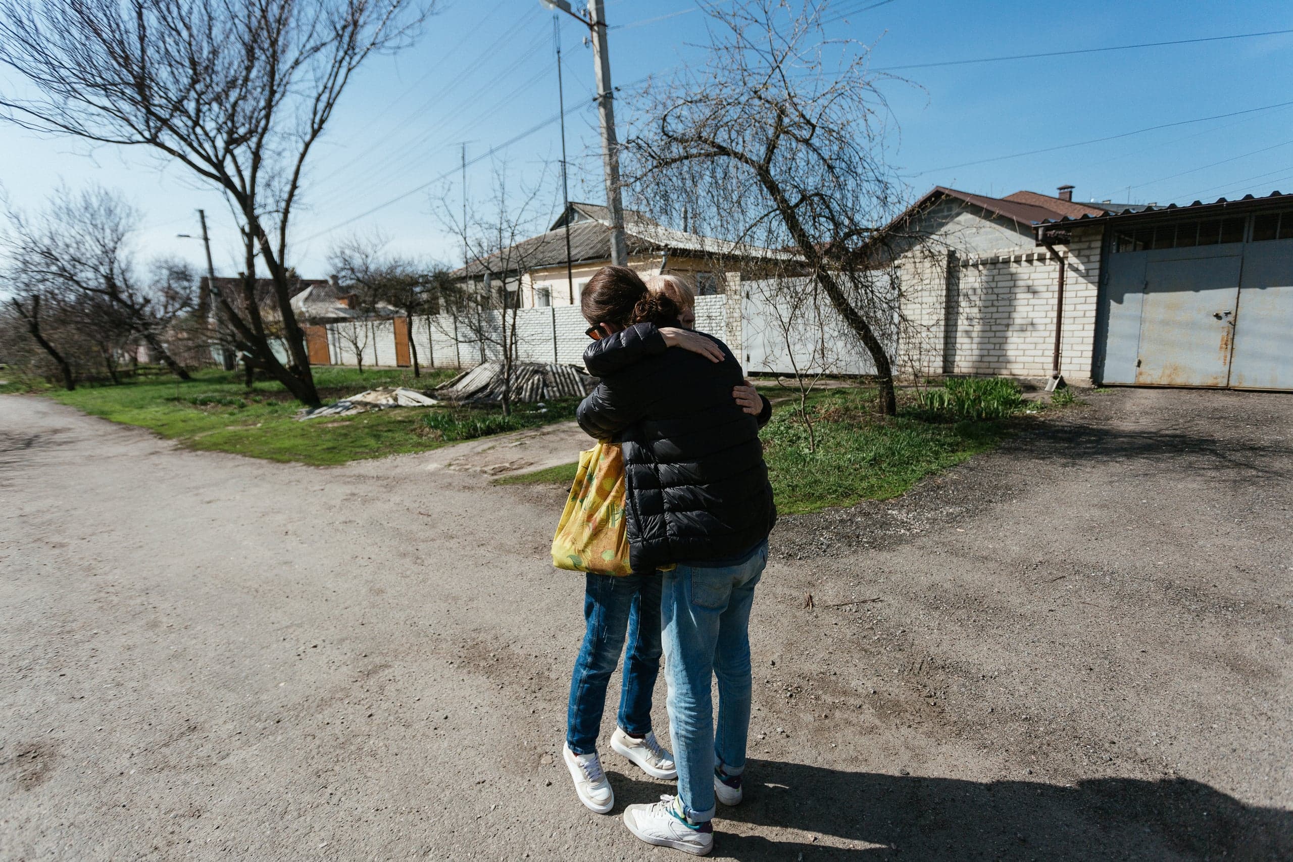 Двое людей в джинсах и куртках обнимаются посреди грунтовой дороги в солнечный день. На заднем плане видны хозяйственные постройки из белого кирпича, металлическая дверь гаража и голые деревья.