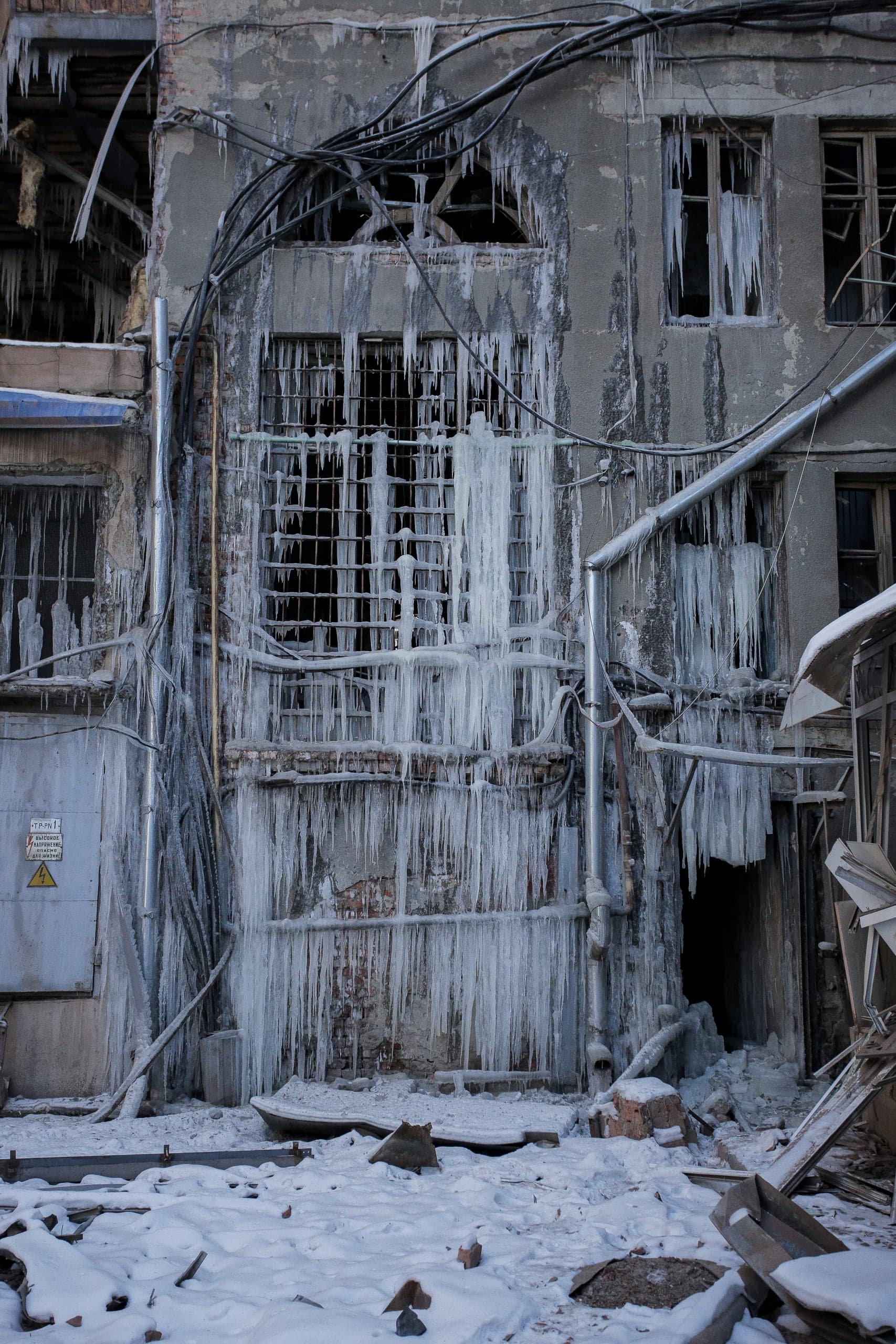 Разрушенный фасад здания, полностью покрытый длинными сосульками, нависающими над окнами, трубами и проводами. На щитке слева: "ОПАСНО", "ЭЛЕКТРИЧЕСТВО".