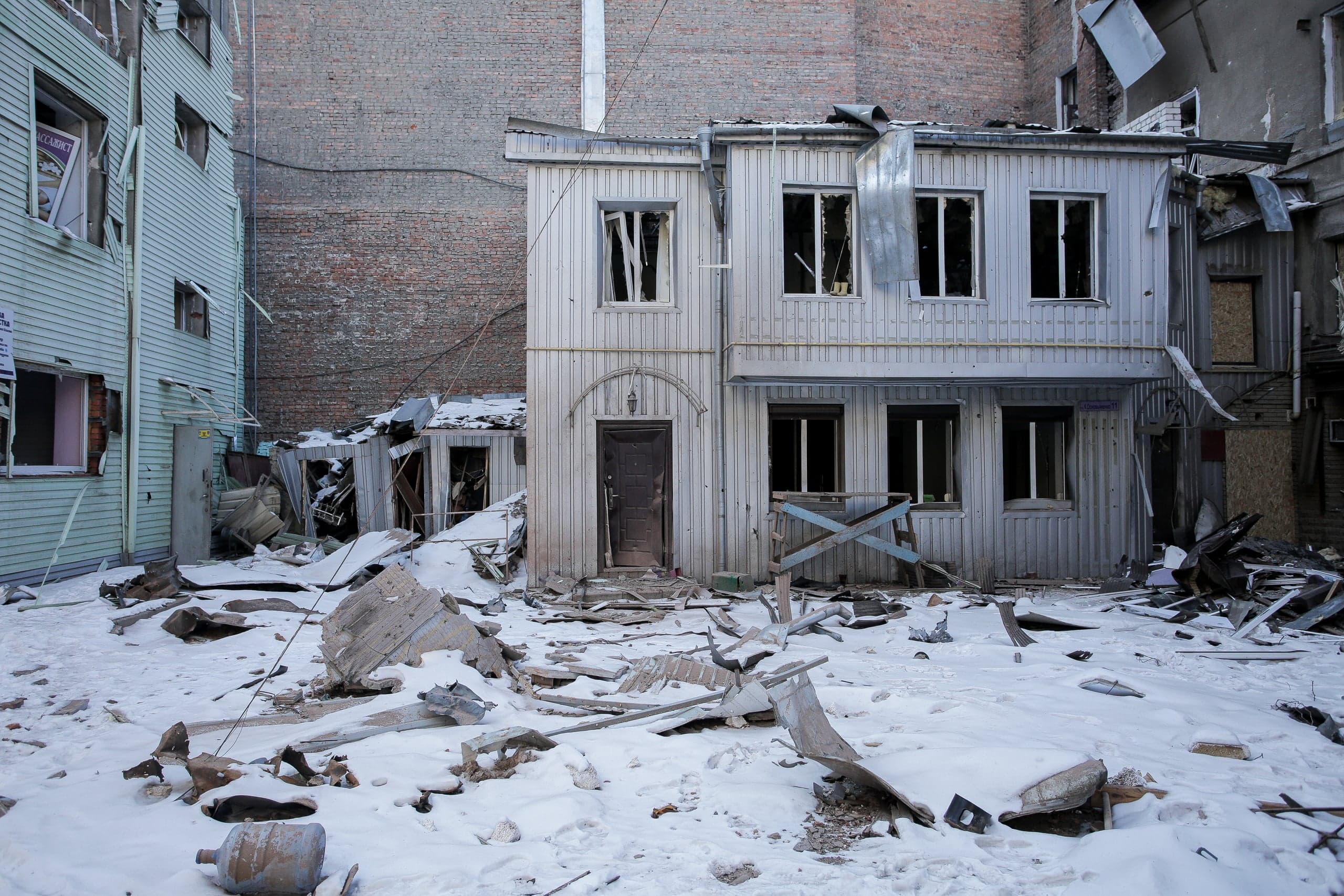 Поврежденное деревянное двухэтажное здание с выбитыми окнами и заснеженный двор, усеянный обломками. В окне виден текст "АССИСТ".