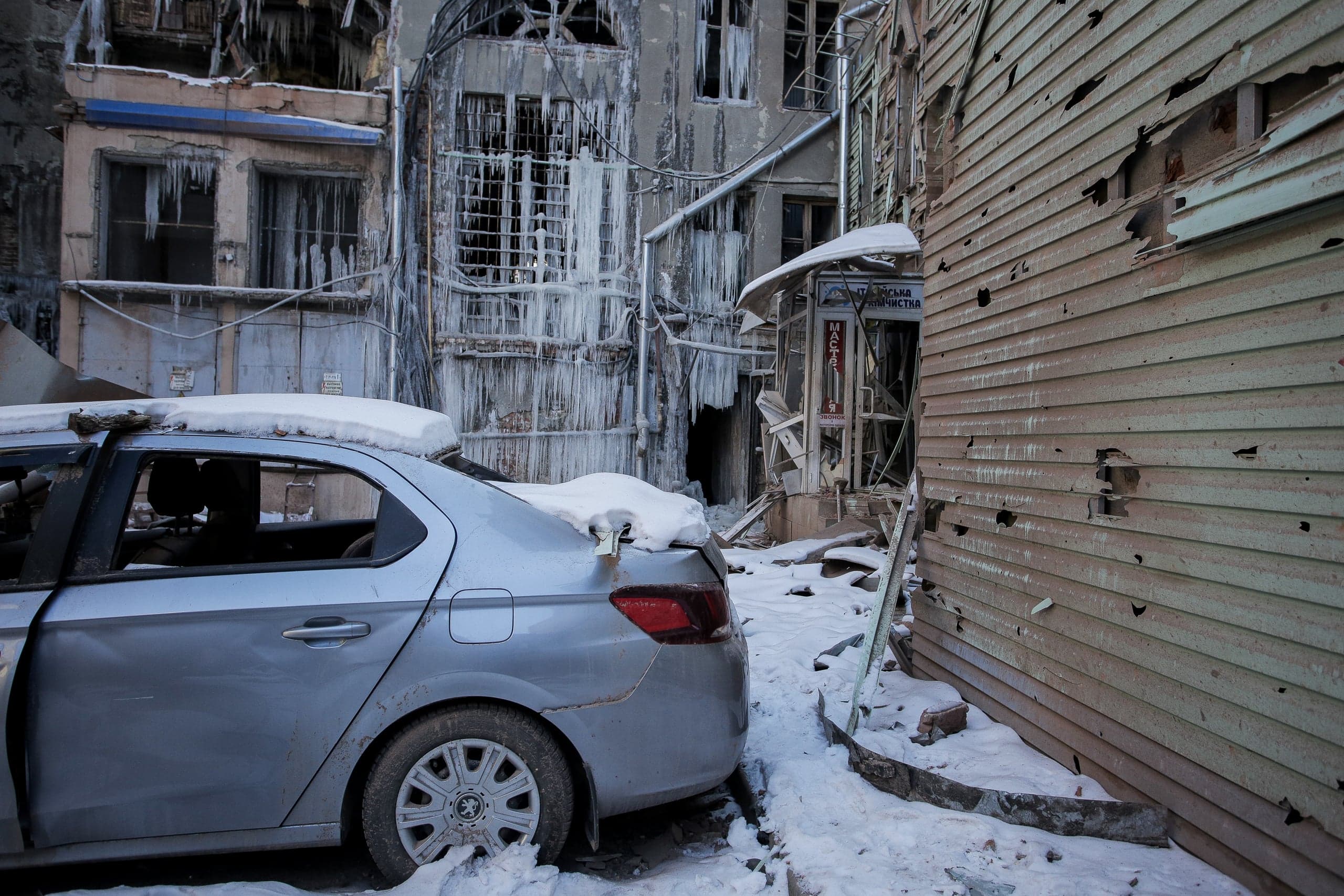 Взорванный автомобиль на фоне разрушенных, покрытых льдом зданий и изрешеченной пулями стены. Вывеска "ХІМЧИСТКА".