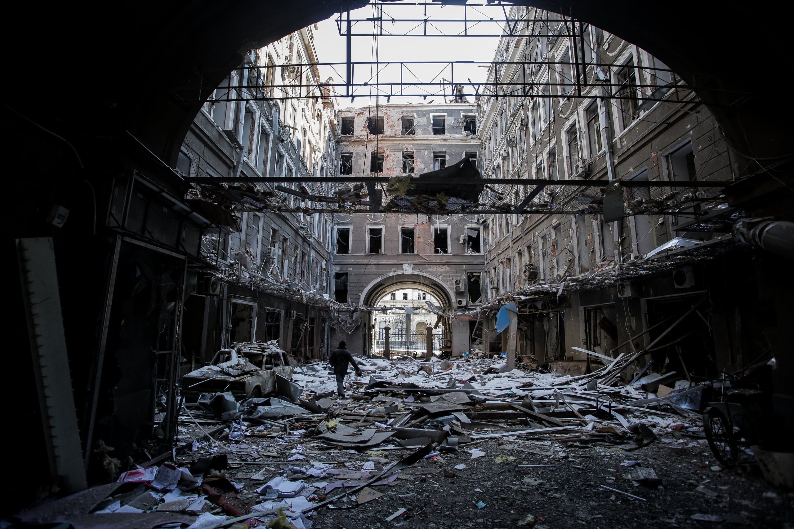 Разрушенный внутренний двор с обломками и поврежденными фасадами зданий. Силуэт человека идет сквозь руины к арке в глубине.