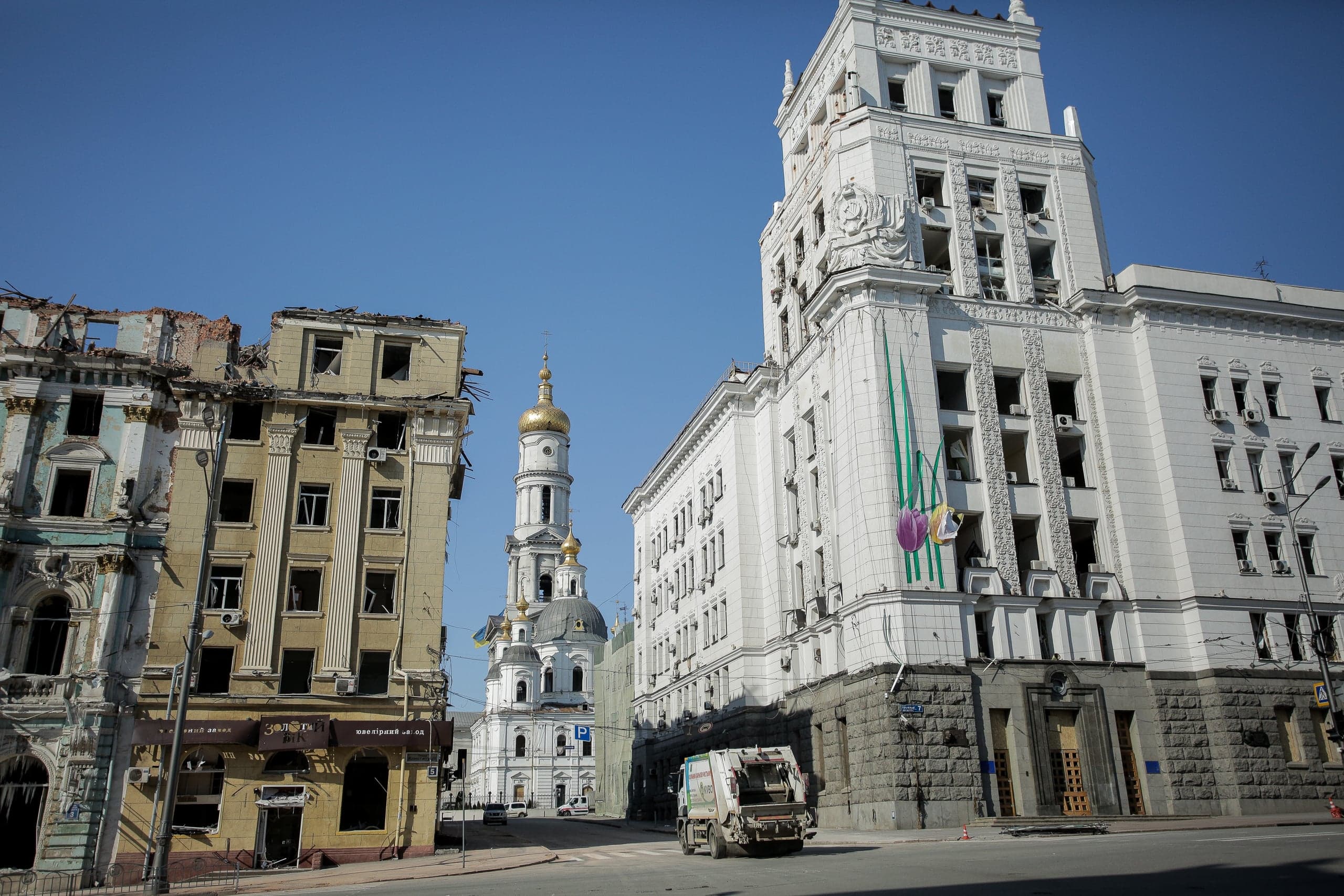 Разрушенные многоэтажные дома и поврежденное высокое белое здание в городе, между ними — церковь с золотым куполом. На улице мусоровоз.