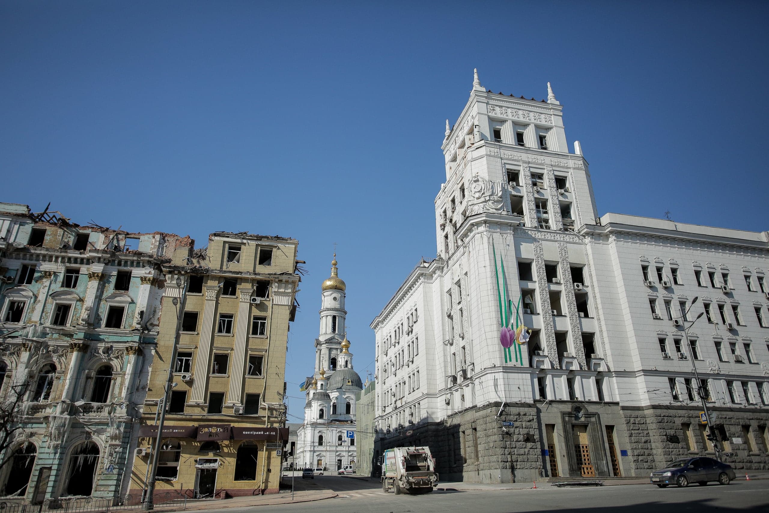 Поврежденные и разрушенные здания на городской улице. Слева — руины жилых домов, справа — большое белое административное здание с выбитыми окнами. В центре виден высокий православный собор с золотым куполом.
