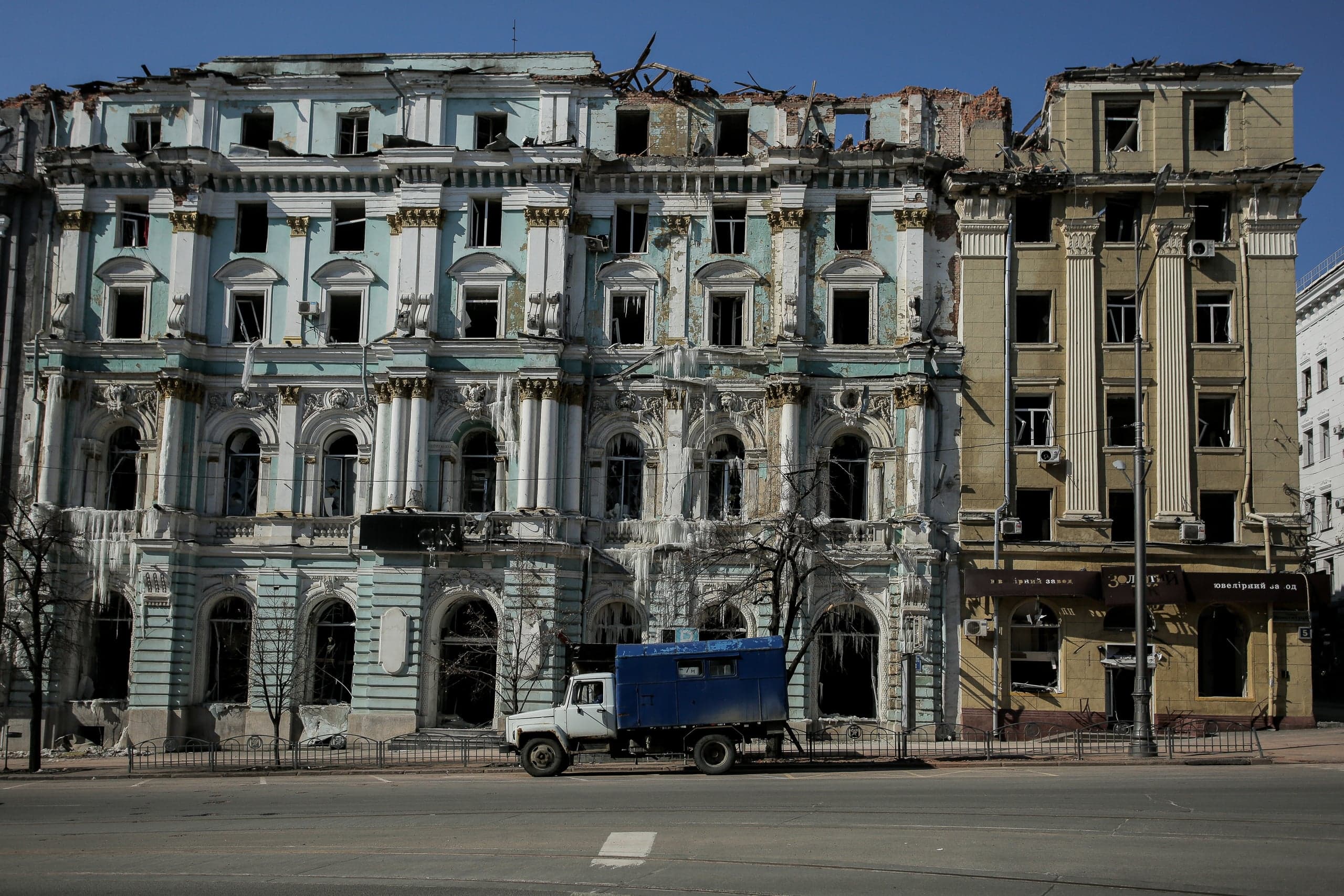 Разрушенные многоэтажные здания (голубое и бежевое) с обрушенными крышами и выбитыми окнами. На улице припаркован грузовик с синим кузовом.
