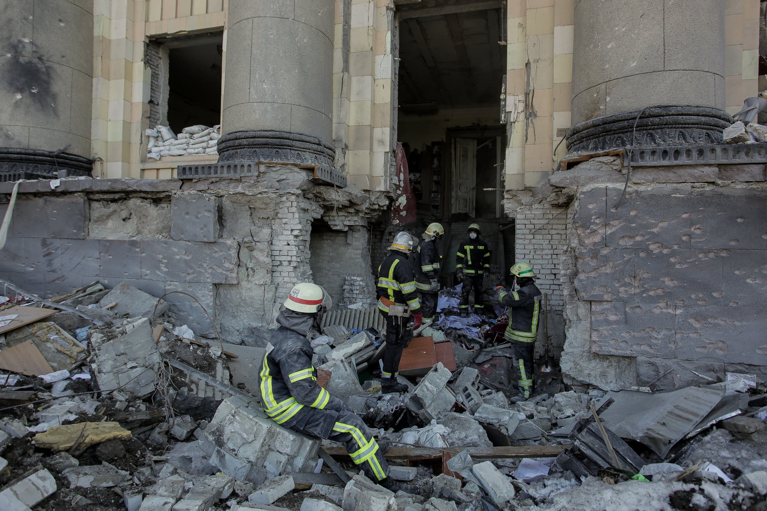 Спасатели работают среди руин разрушенного здания с колоннами, вокруг много обломков и мусора.