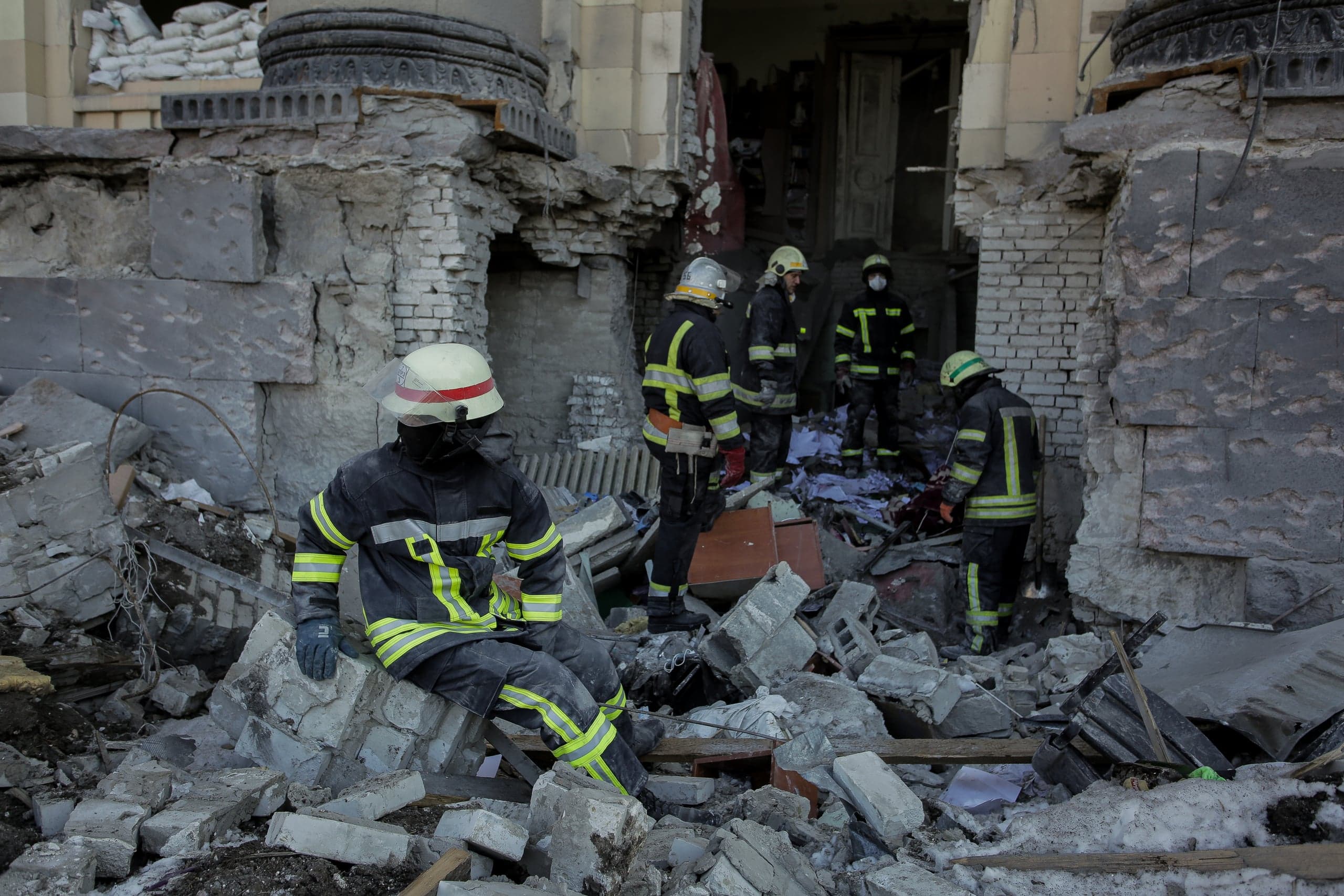 Пожарные среди обломков разрушенного здания; один спасатель сидит на куче кирпичей на переднем плане.