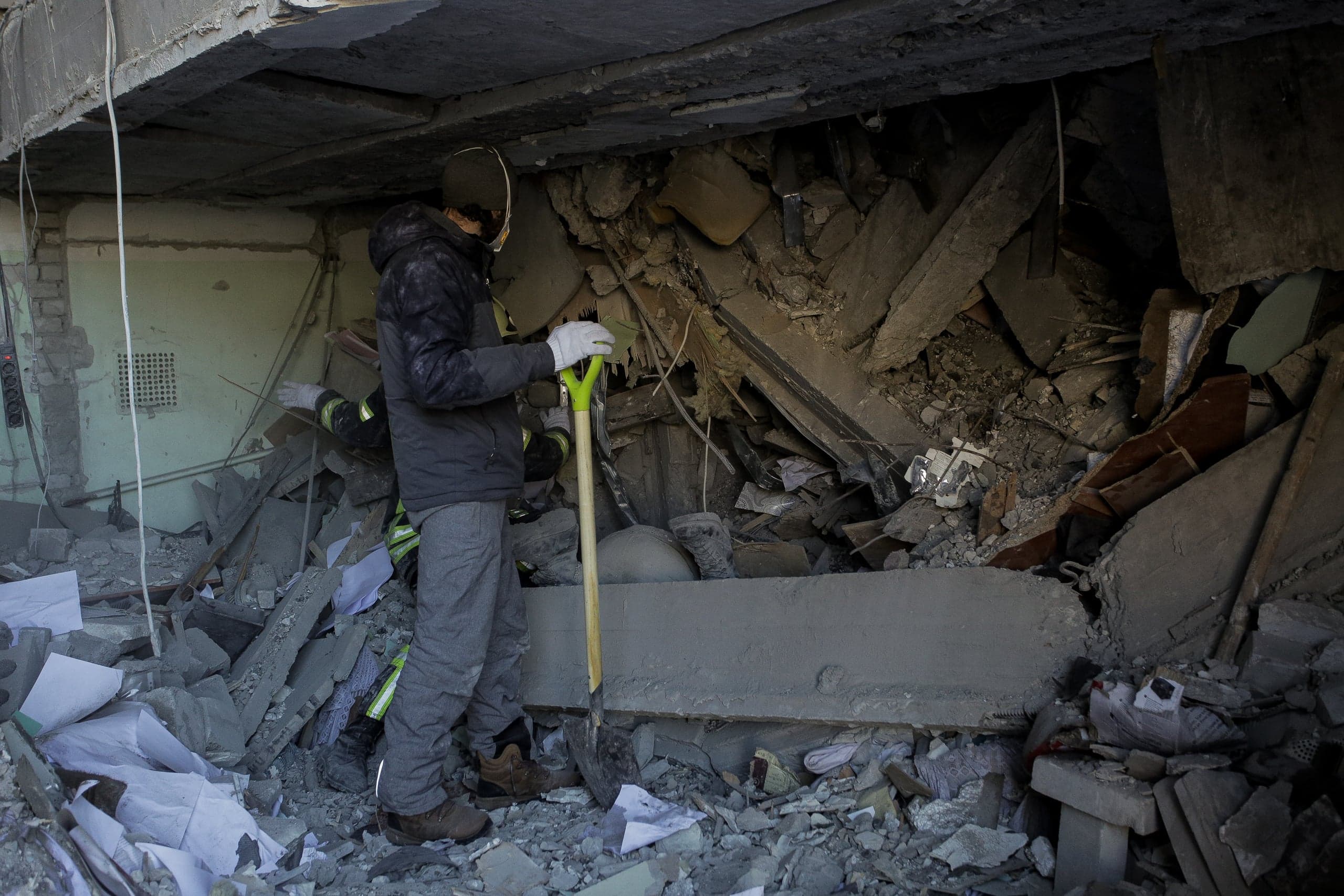 Спасатель с лопатой стоит среди обломков полностью разрушенного здания.