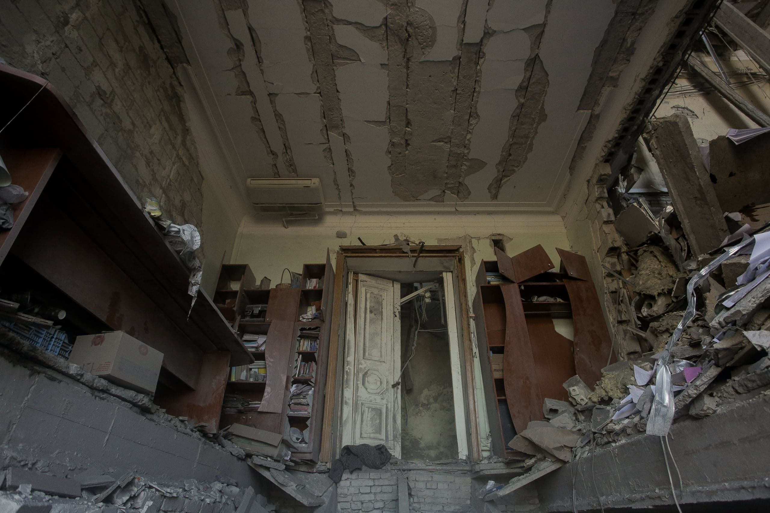 Разрушенное помещение библиотеки или офиса с поврежденным потолком, обломками бетона и покосившейся мебелью.