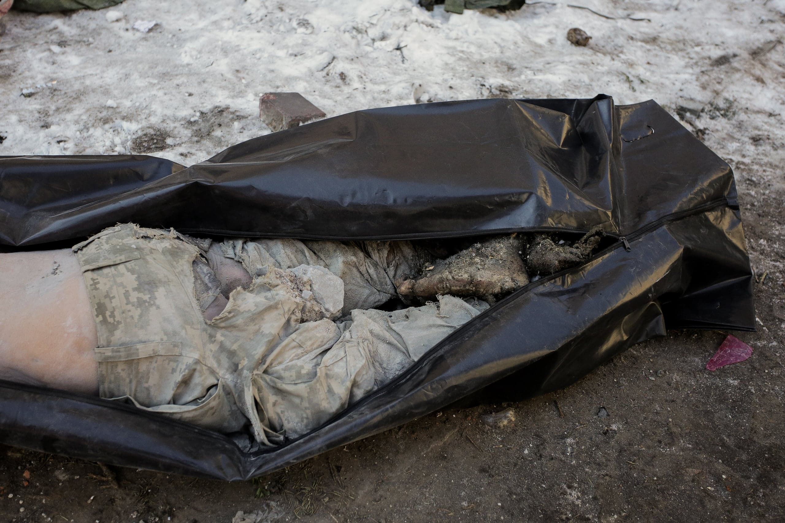 Частично открытый черный мешок с телом в камуфляжной форме на грязном снегу.