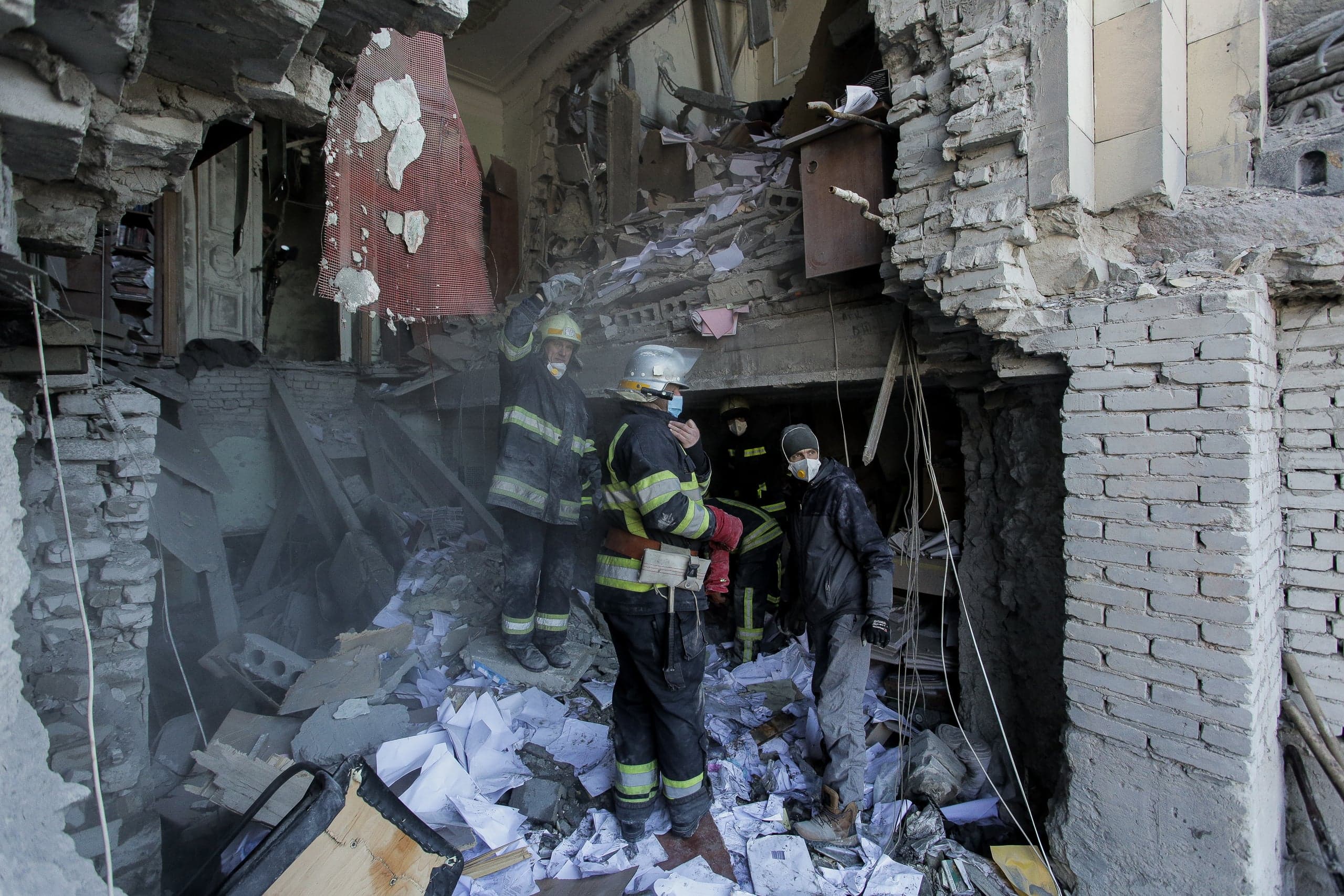 Спасатели и гражданские в масках работают среди обломков и бумаг в разрушенном здании.
