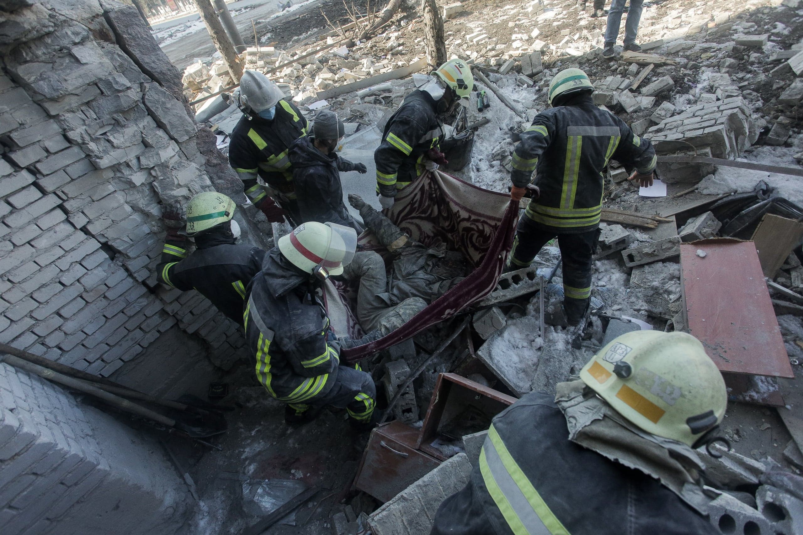 Спасатели извлекают тело, завернутое в ковер, из-под завалов разрушенного здания.