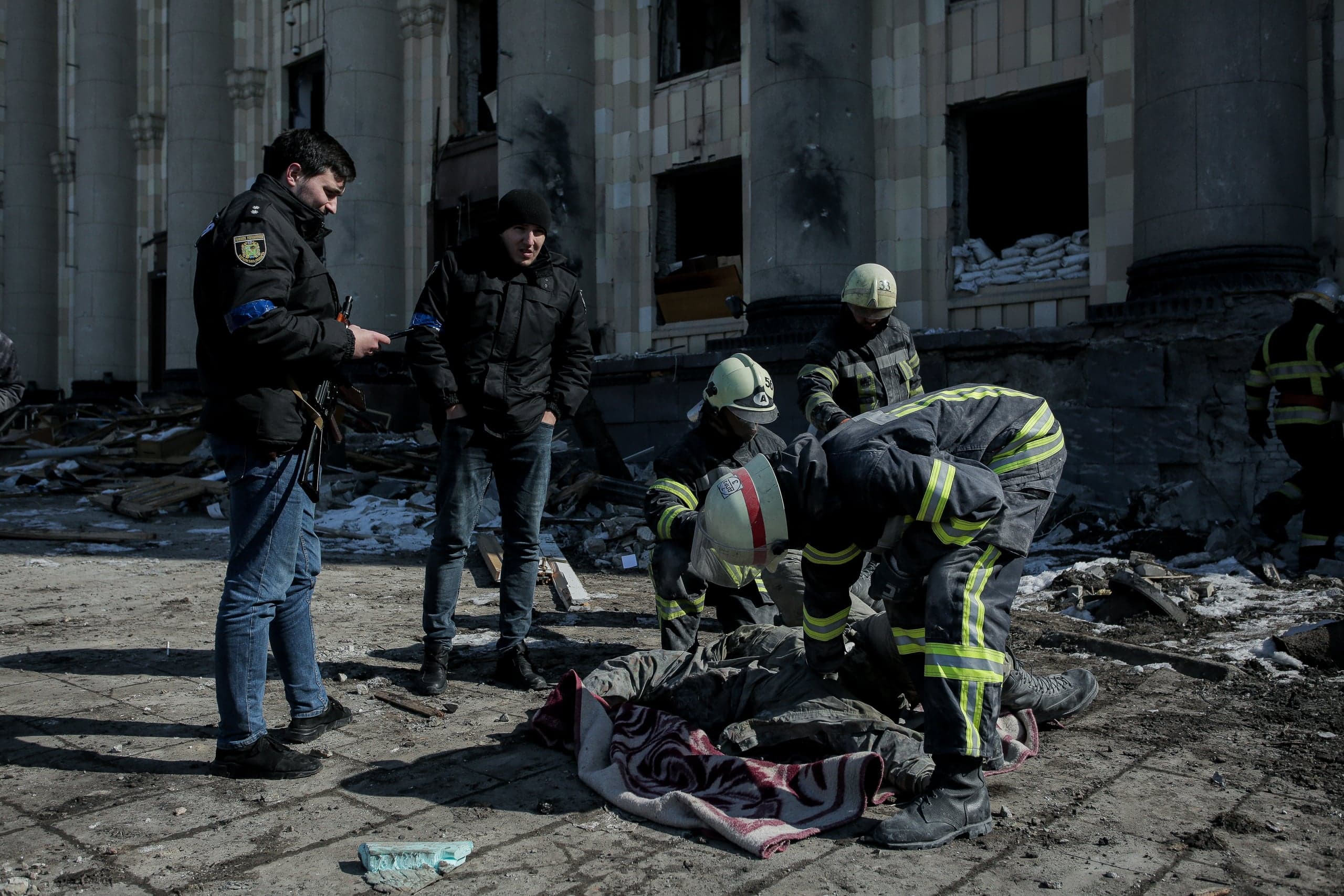 Спасатели и полиция возле разрушенного здания, пожарные склонились над телом на земле, покрытым пледом.