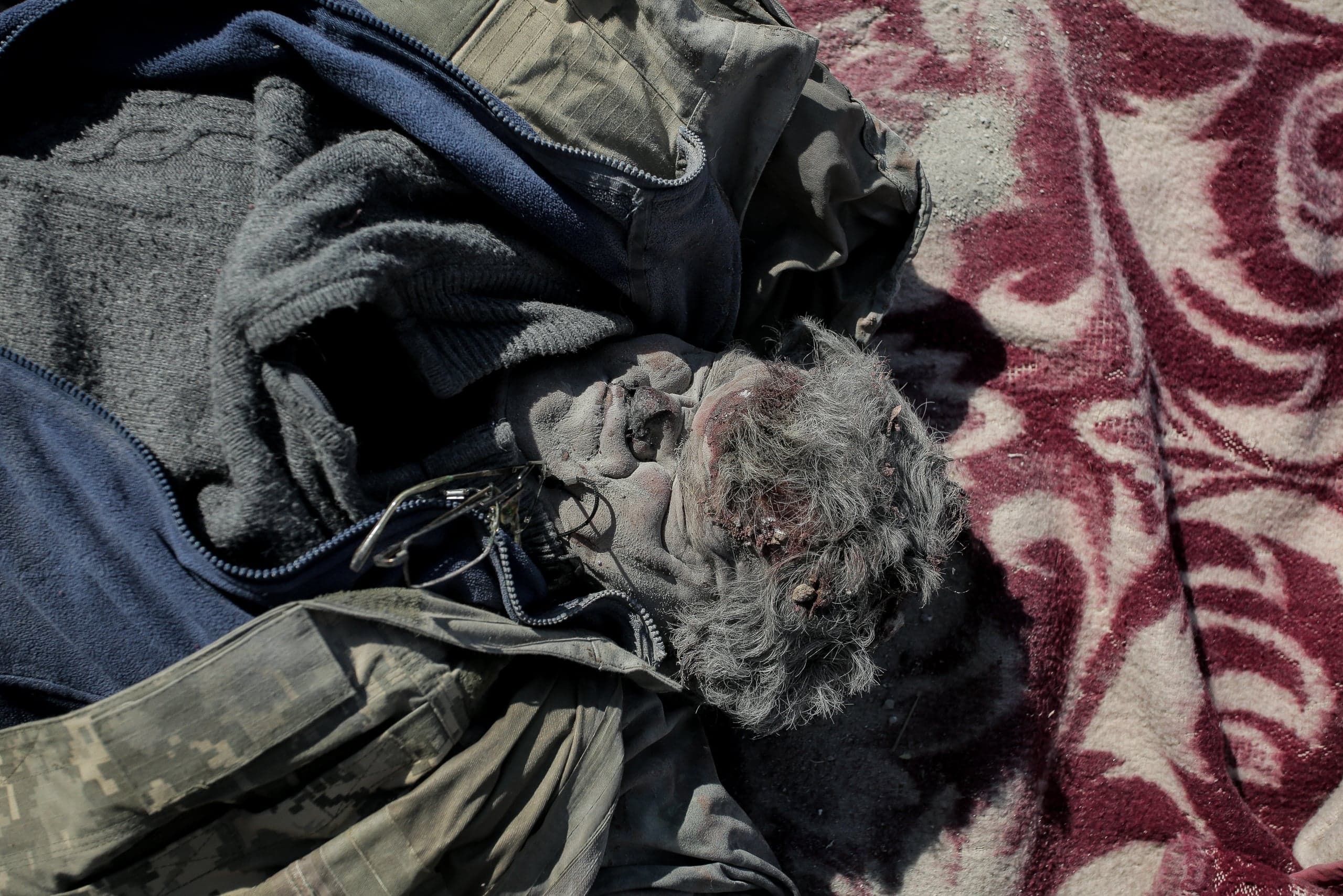Останки человека в одежде и очках, покрытые пылью, на бордовом узорчатом одеяле.