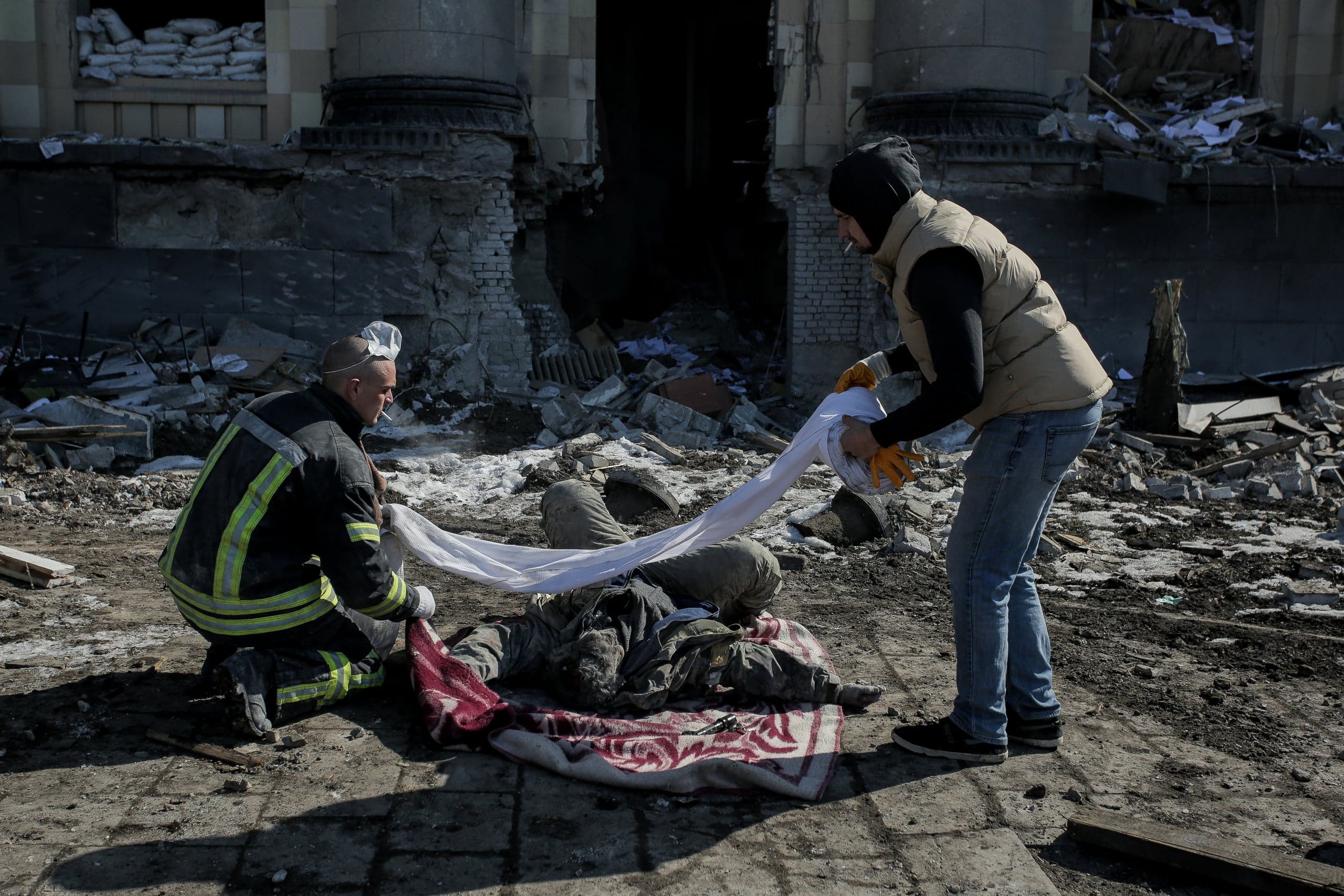 Спасатель и гражданский накрывают белой тканью тело погибшего на фоне разрушенного обстрелом здания.