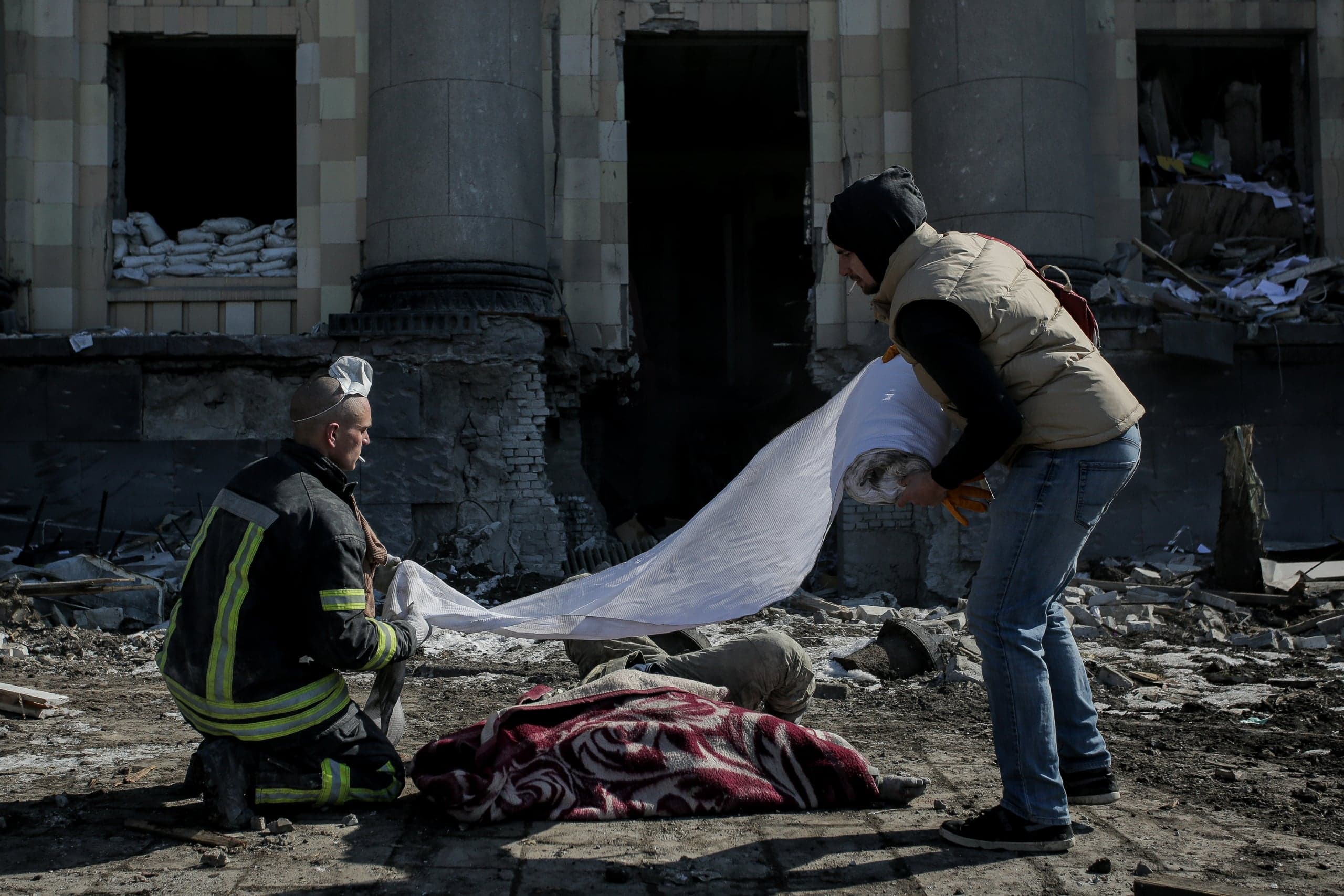 Пожарный и гражданский стоят над телом, накрытым одеялом, на фоне разрушенного здания с колоннами. Мужчины разворачивают белое полотно.