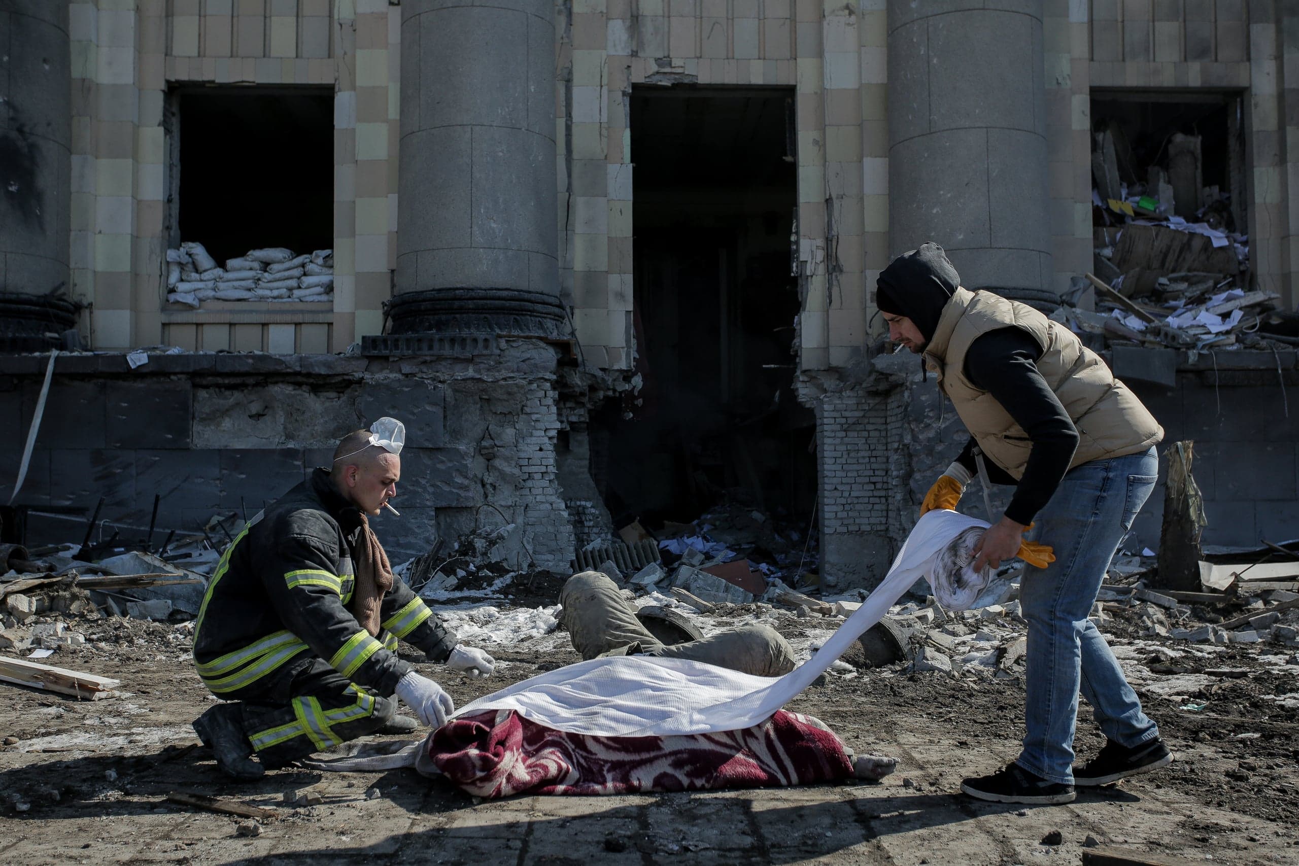 Спасатель и гражданский оборачивают тело погибшего одеялом на фоне разрушенного здания.