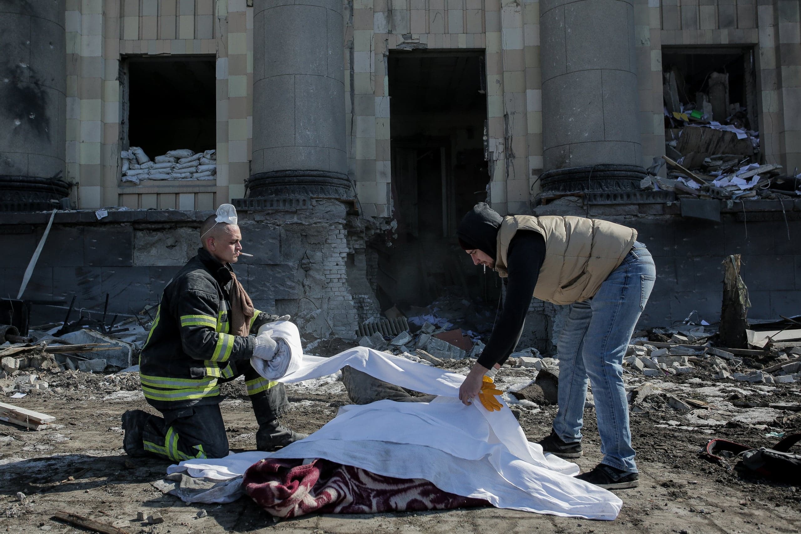 Спасательные работы среди руин: двое мужчин накрывают тело белой тканью перед сильно поврежденным зданием с колоннами.