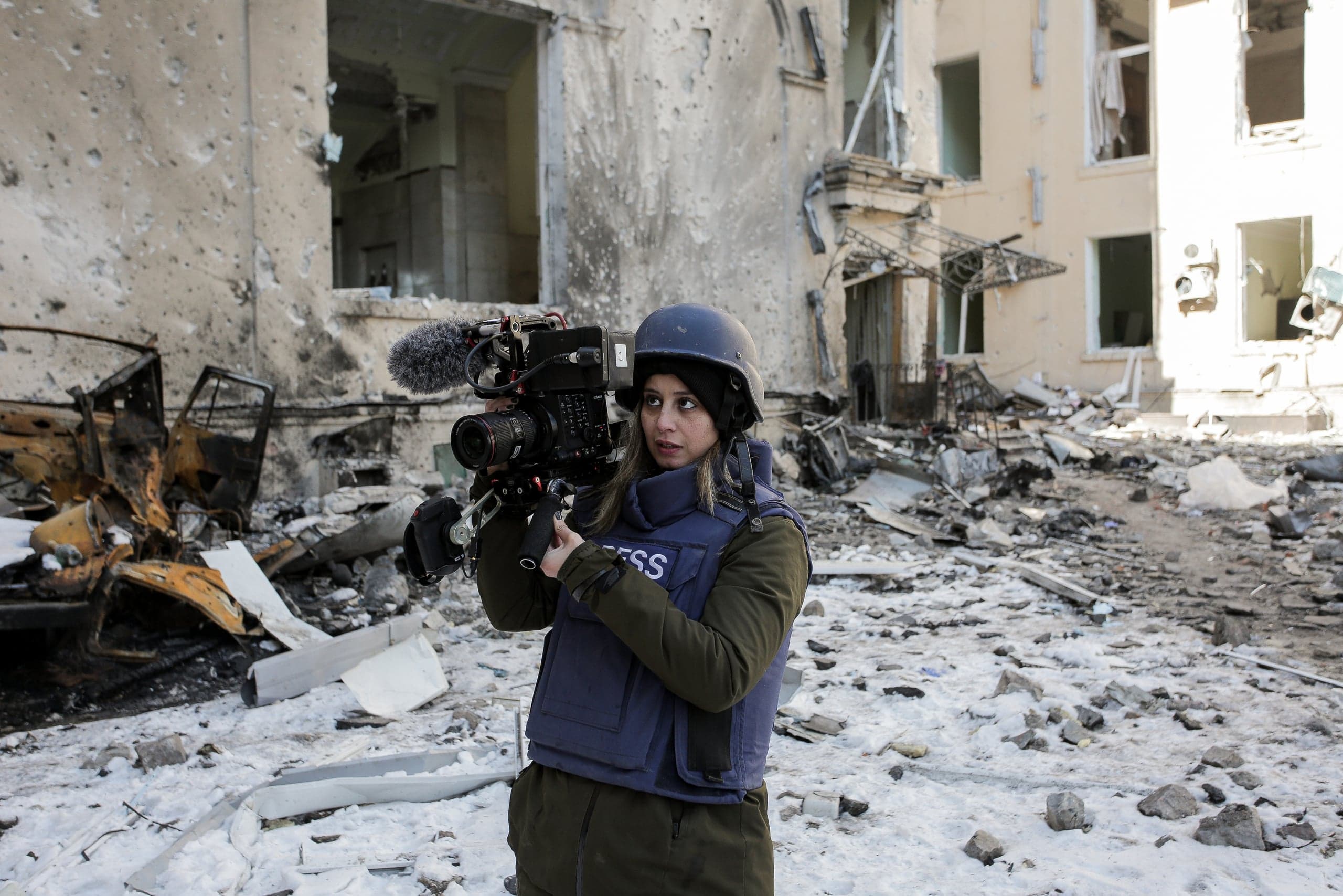 Военный корреспондент в шлеме и бронежилете с надписью "PRESS" снимает разрушенное здание на профессиональную видеокамеру среди обломков и снега.