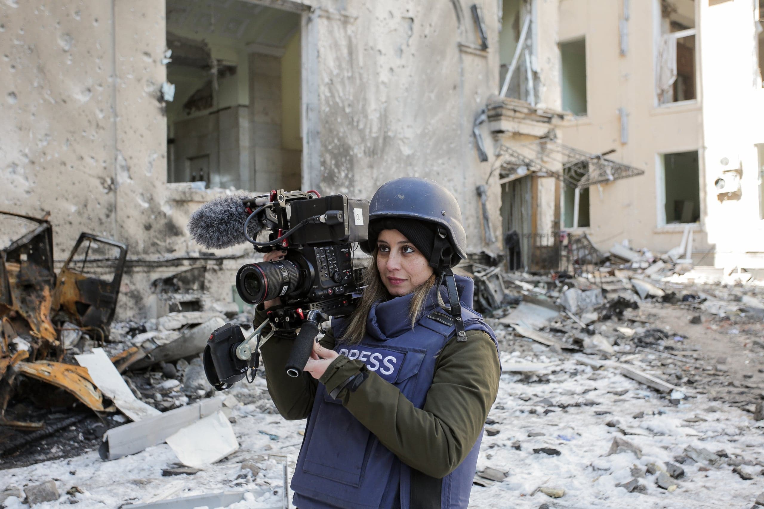 Женщина-оператор в каске и бронежилете "PRESS" снимает на камеру разрушенное здание и обломки.