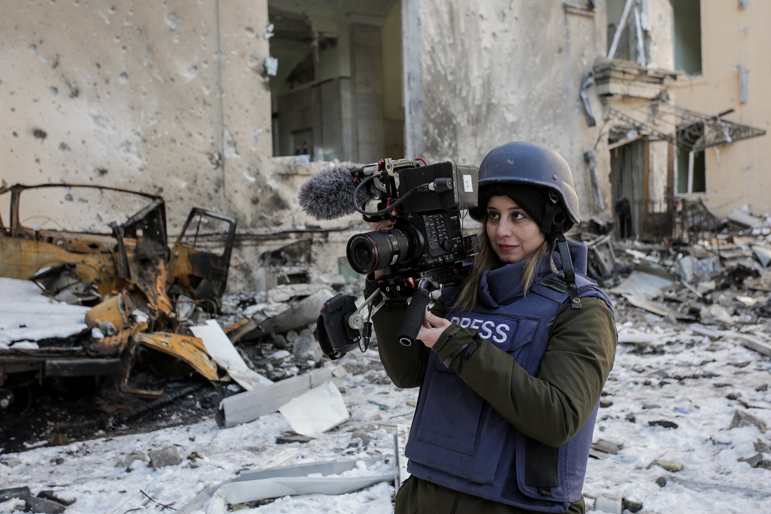Видеооператор в шлеме и бронежилете с надписью "PRESS" снимает репортаж среди руин разрушенного здания и сгоревшего автомобиля.
