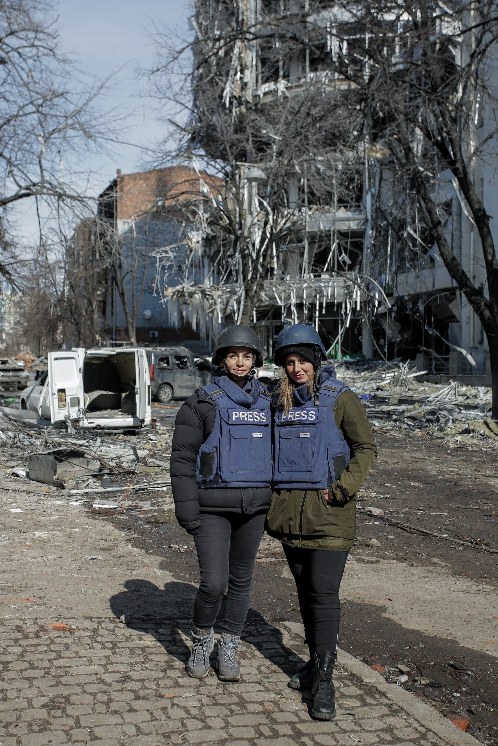 Две женщины-журналистки в бронежилетах с надписью "PRESS" и касках стоят на фоне разрушенного здания и обломков.