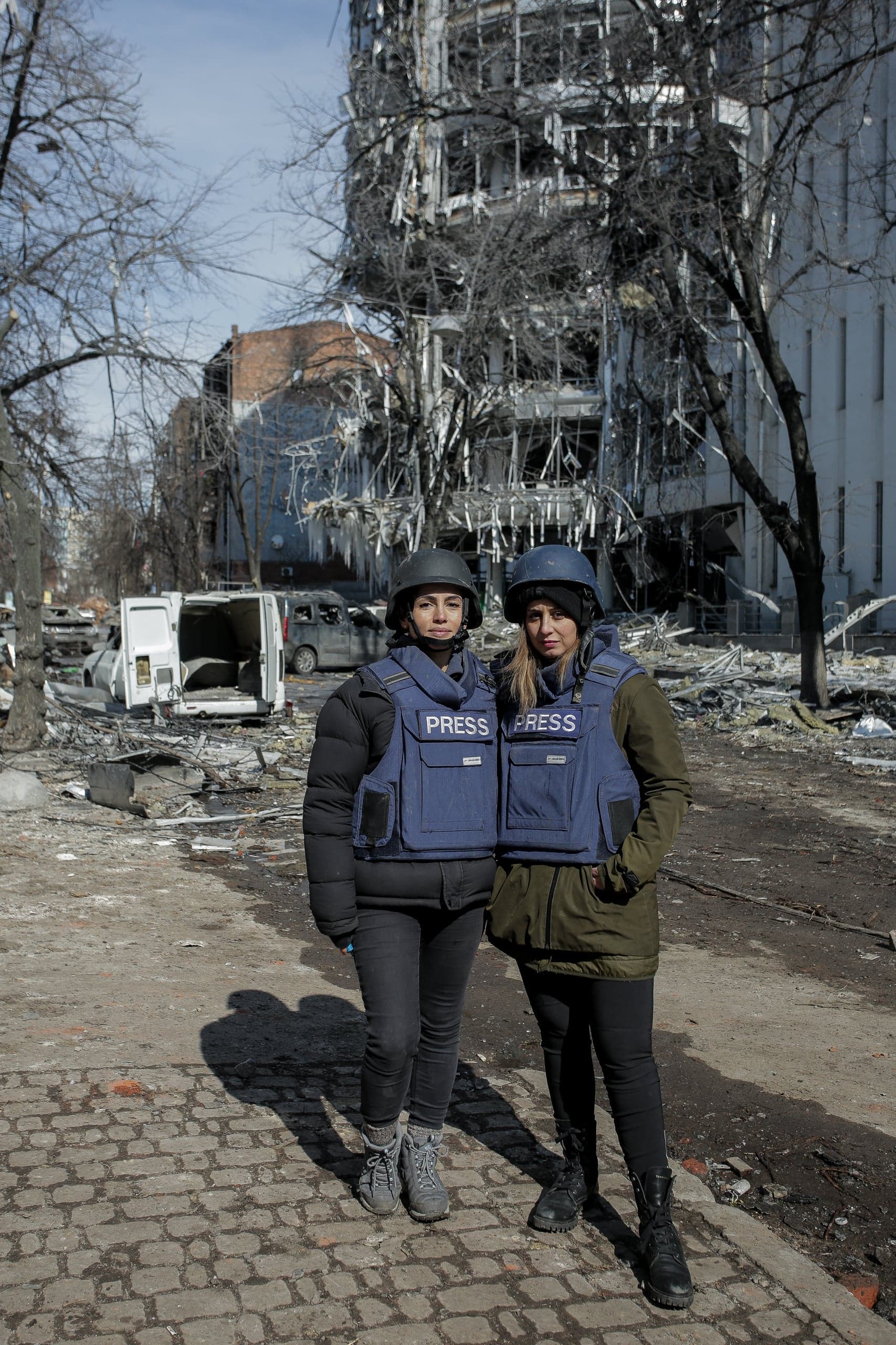 Две журналистки в шлемах и бронежилетах "PRESS" на фоне разрушенного здания.