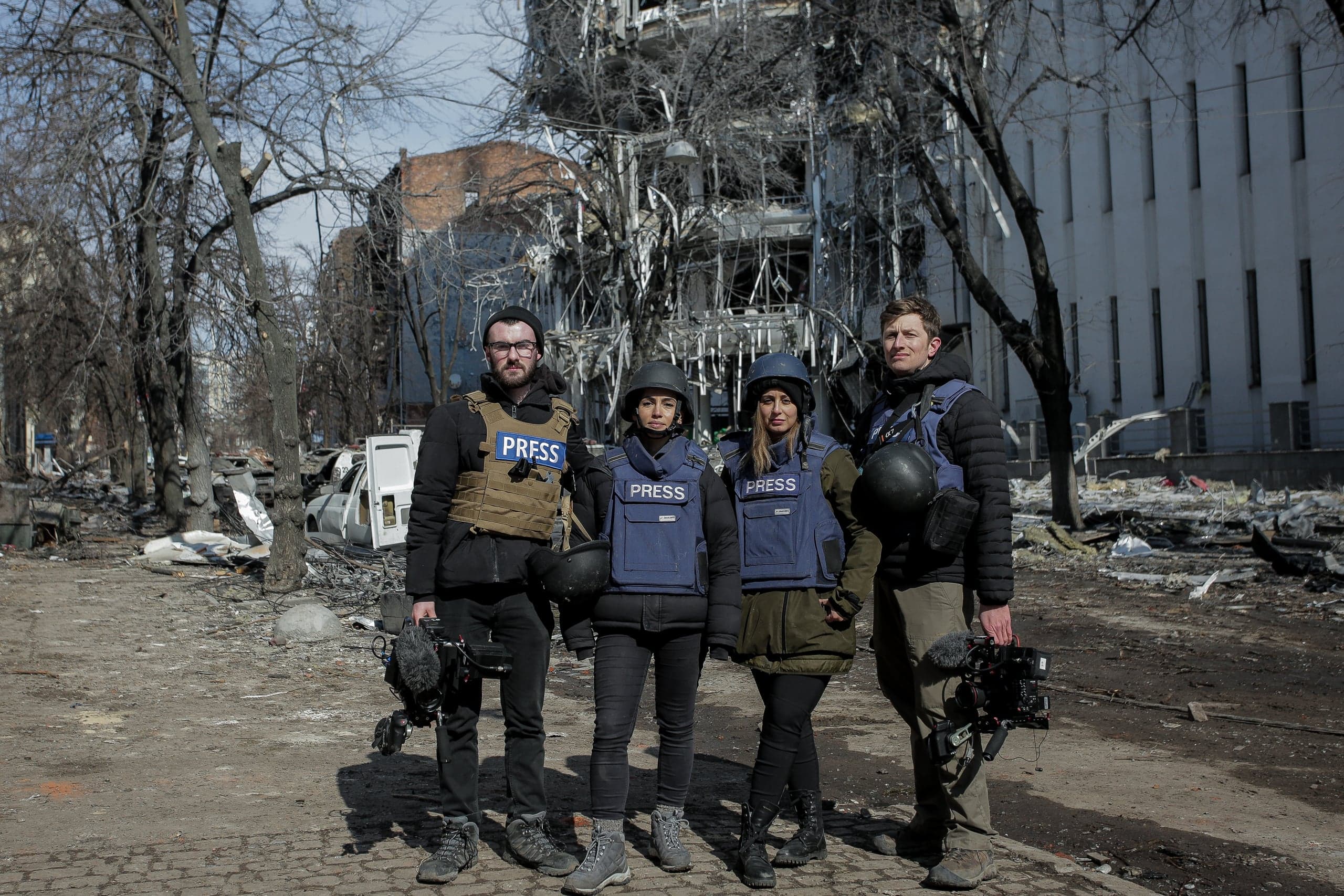 Четыре журналиста в бронежилетах с надписью "PRESS" стоят на разрушенной улице на фоне сильно поврежденного здания.