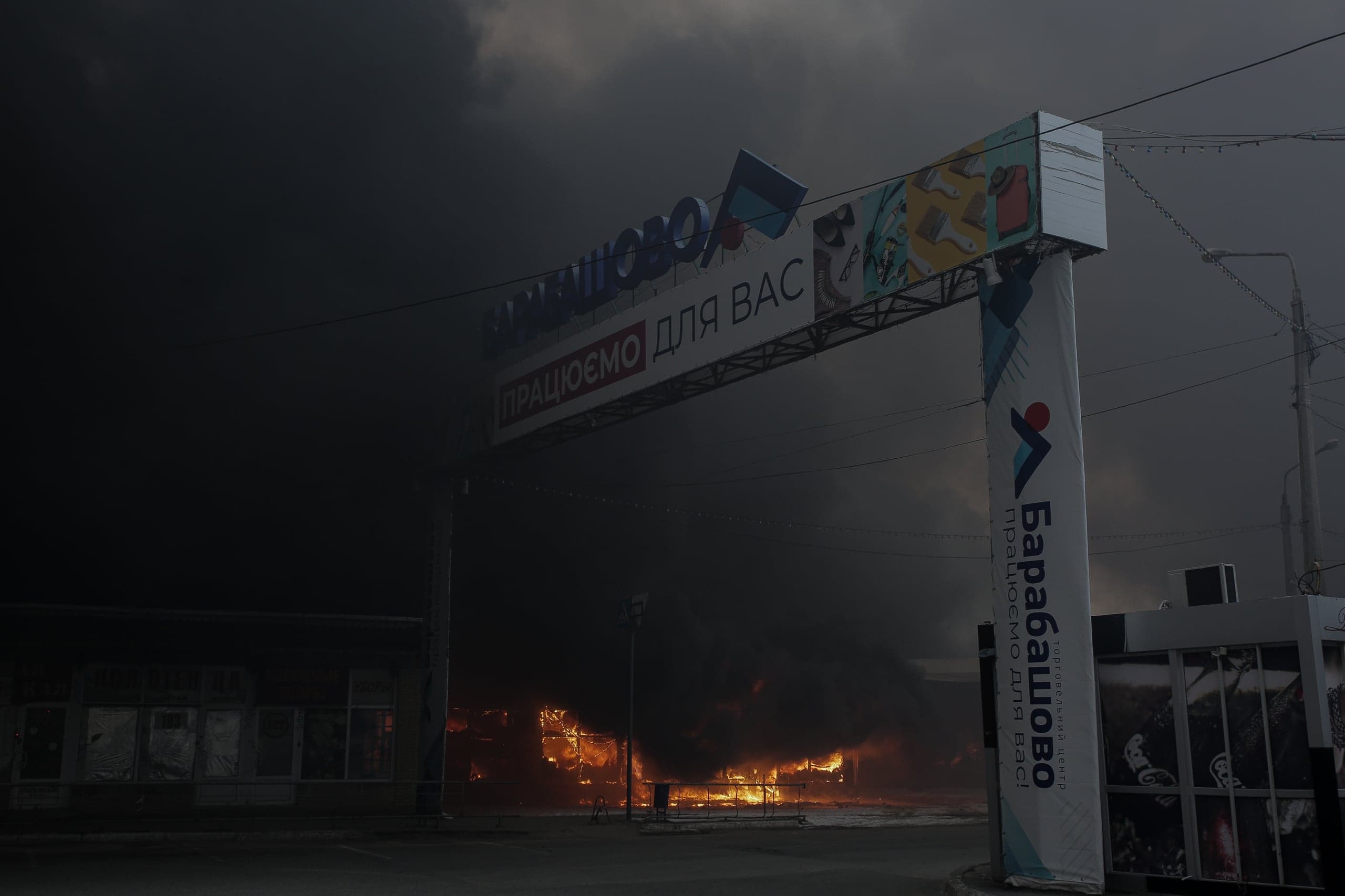 Въездная арка рынка "Барабашово" на фоне мощного пожара и густого черного дыма. Текст: "БАРАБАШОВО", "ПРАЦЮЄМО ДЛЯ ВАС".