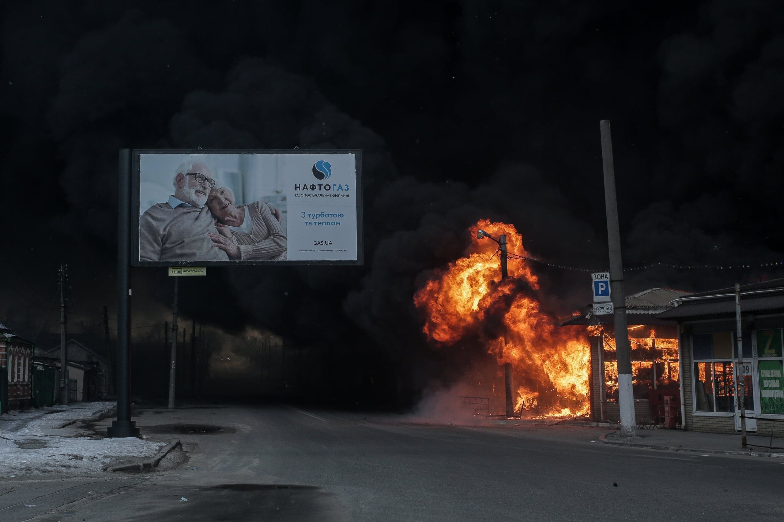 Ночная сцена: яркий огонь и черный дым над горящим зданием рядом с билбордом с рекламой "НАФТОГАЗ" и текстом "З турботою та теплом".