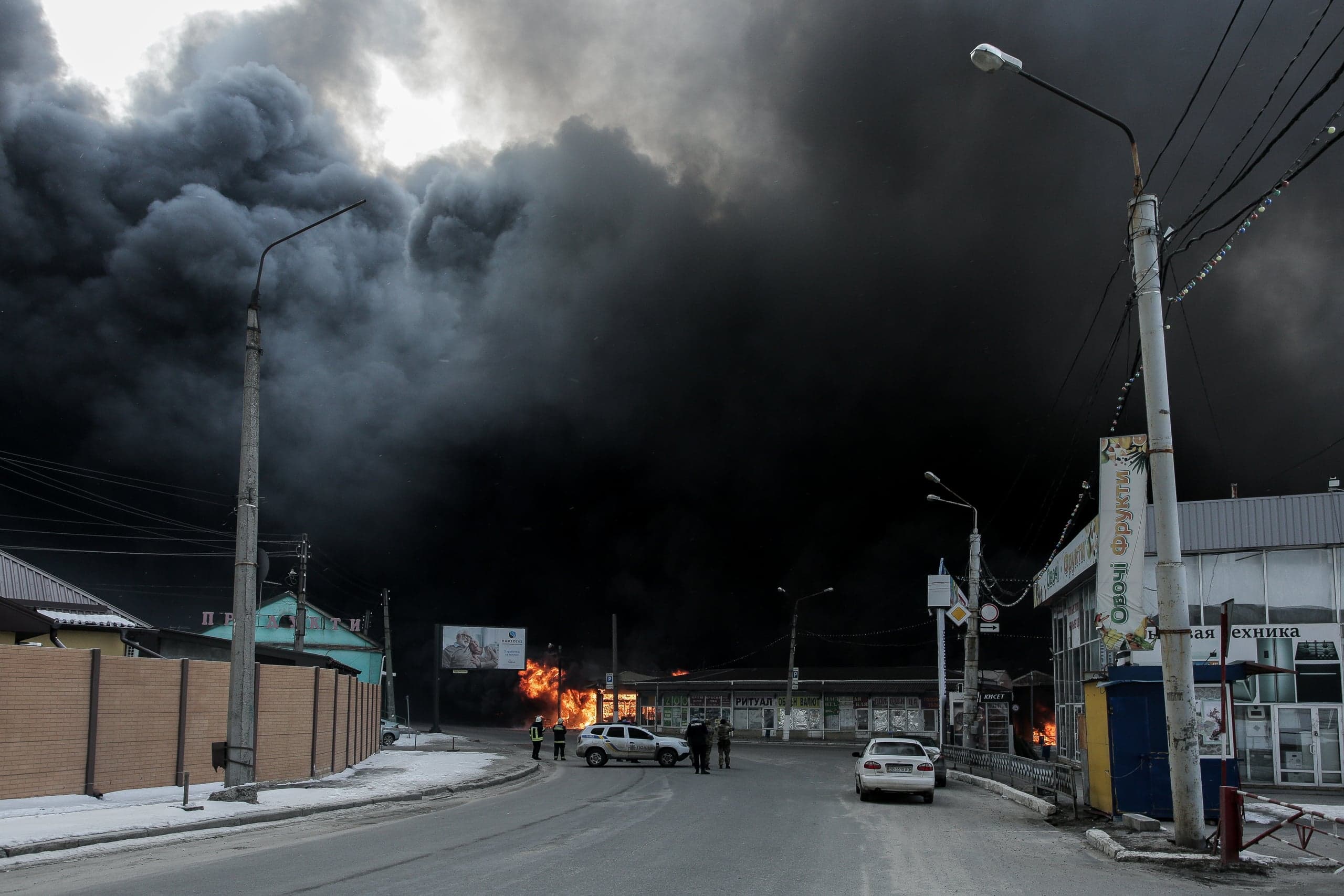 Уличный пожар с массивным столбом густого черного дыма, полностью закрывающим небо. На дороге стоят люди и автомобили; рядом магазин с вывеской "Овочі фрукти".