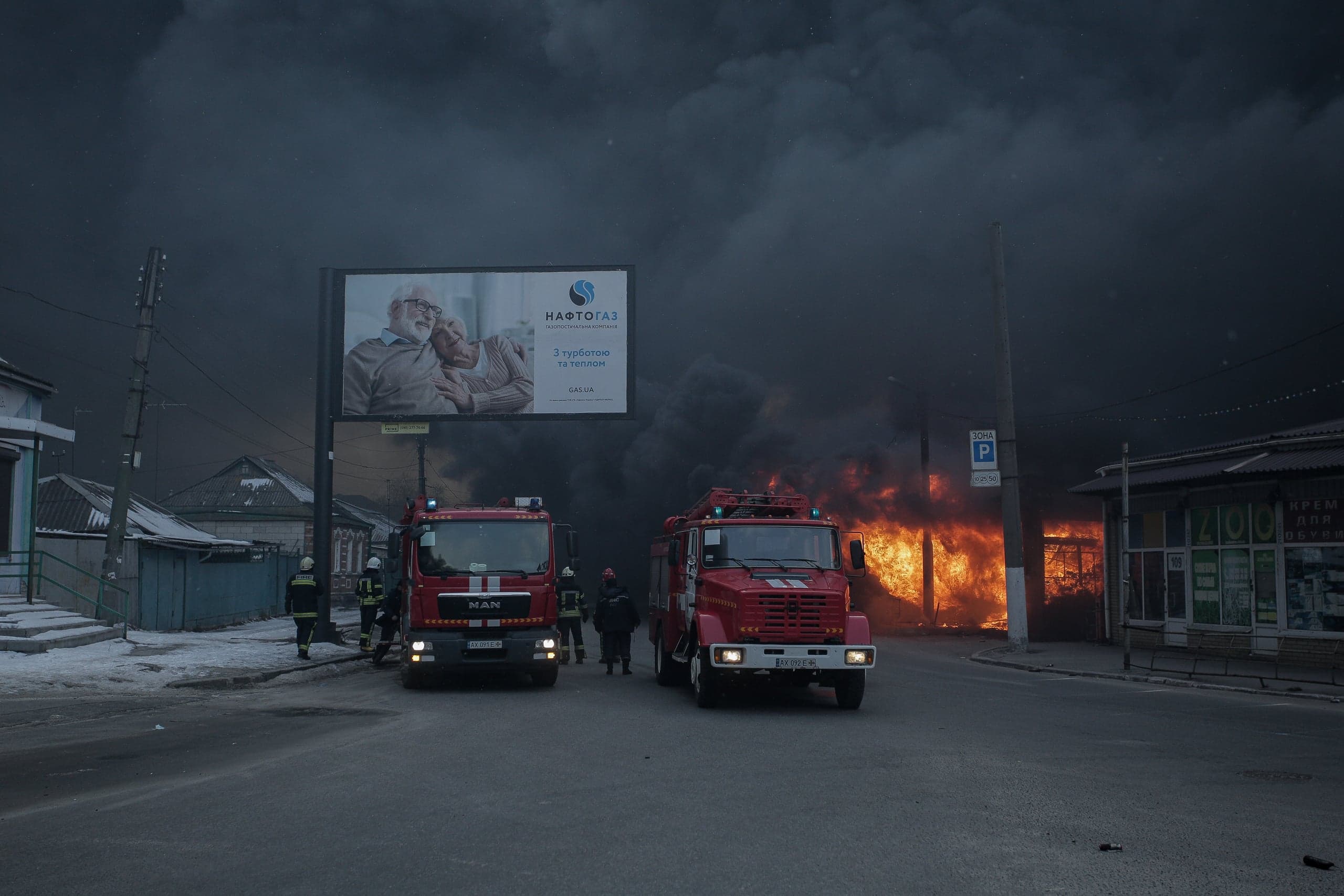 Два пожарных автомобиля и спасатели на улице перед сильным пожаром. Небо затянуто густым черным дымом. На билборде над машинами виден текст "НАФТОГАЗ".