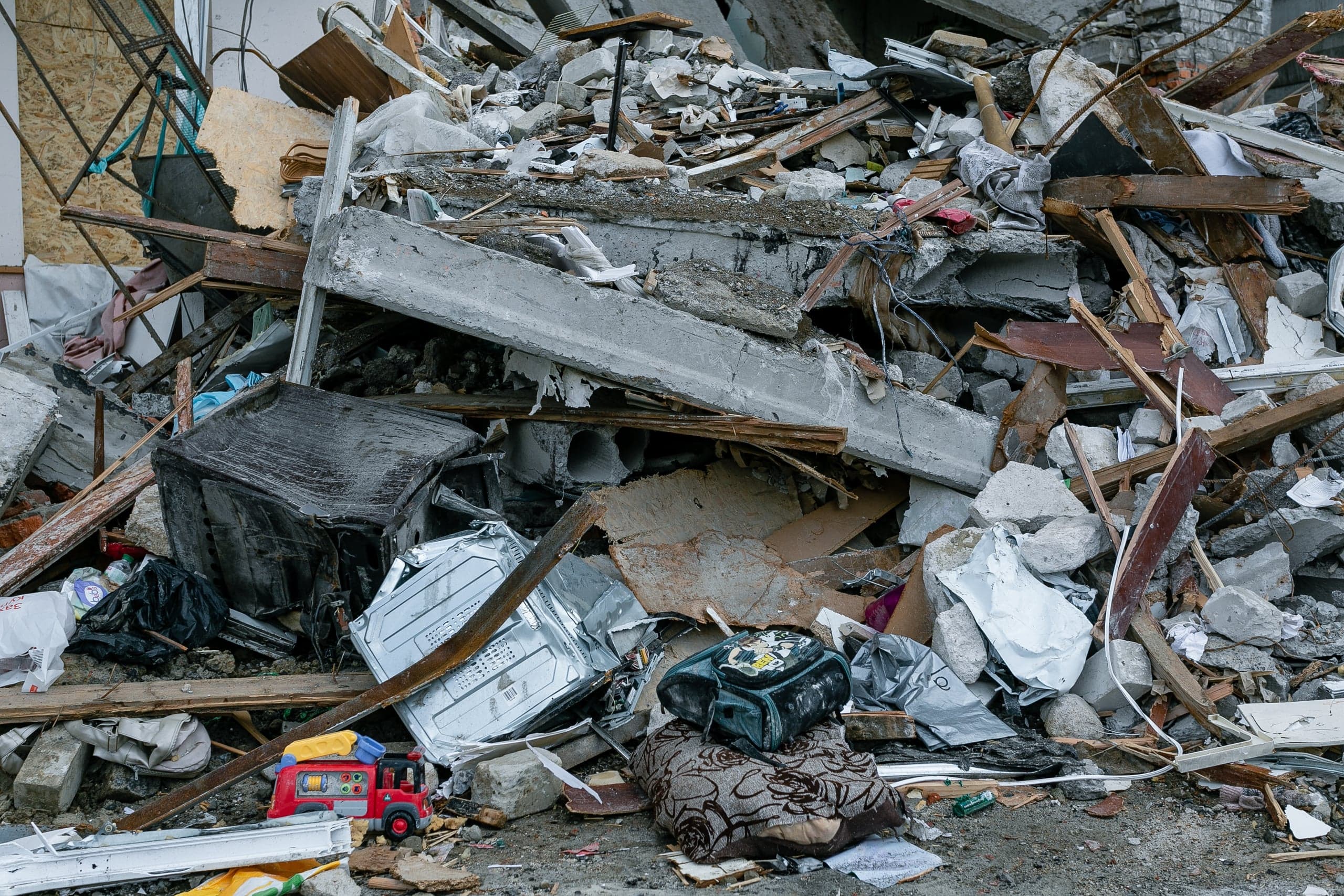 Завал из обломков разрушенного здания: бетонные плиты, деревянные балки, бытовая техника (холодильник, стиральная машина), детская пожарная машинка и рюкзак на подушке.