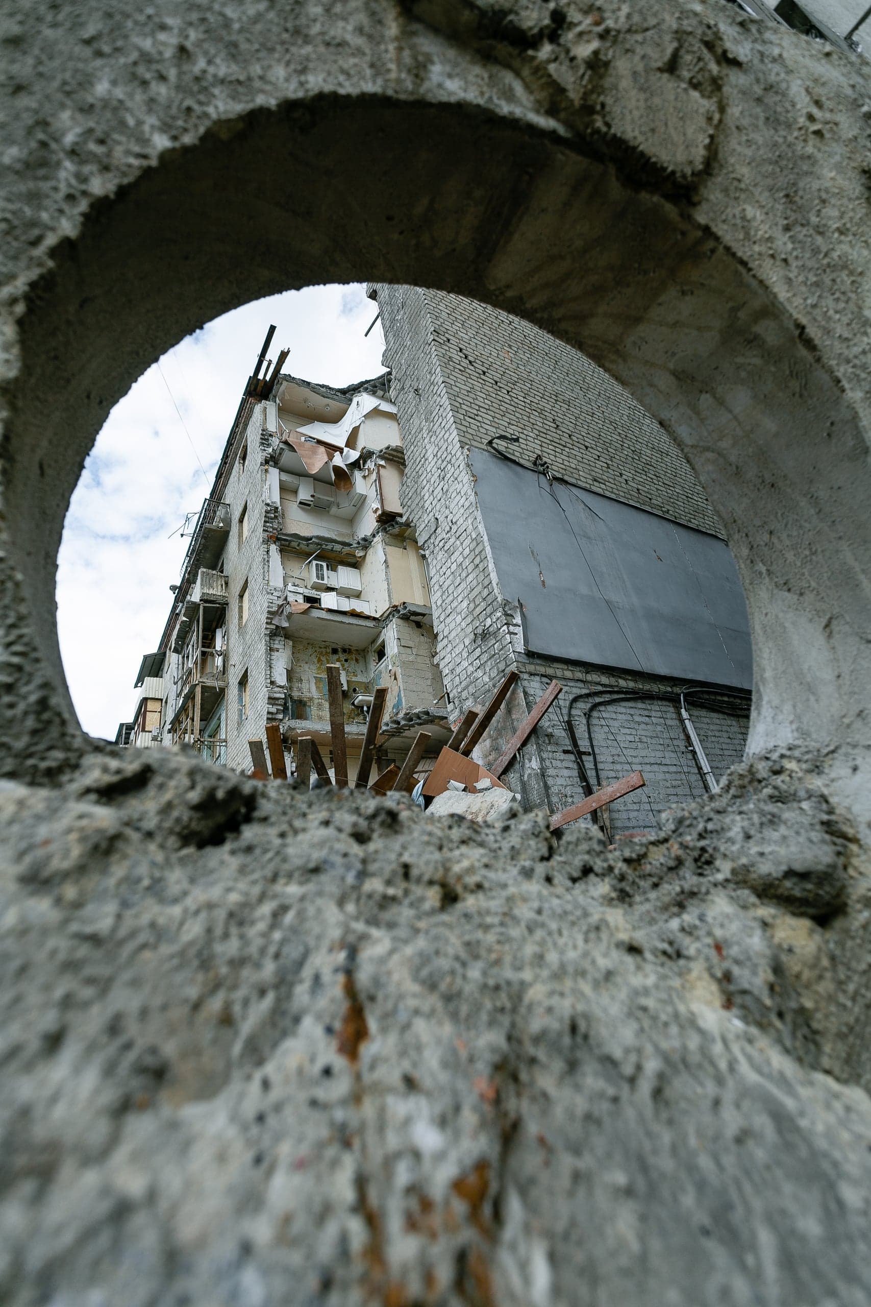 Разрушенный жилой дом, видимый сквозь круглое отверстие в крупнозернистой бетонной стене на переднем плане.
