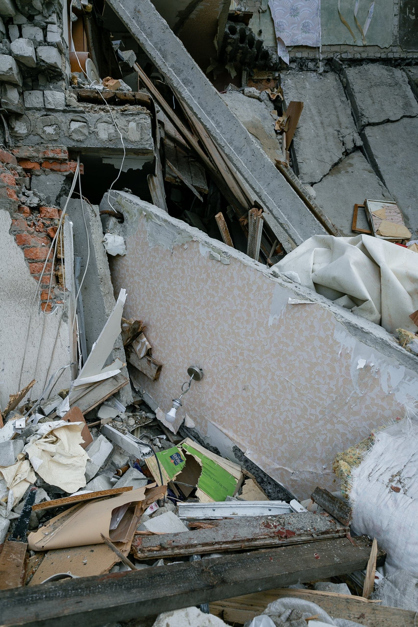 Крупный план обломков разрушенного здания: бетонные плиты, кирпич, фрагмент стены с обоями и свисающим светильником.