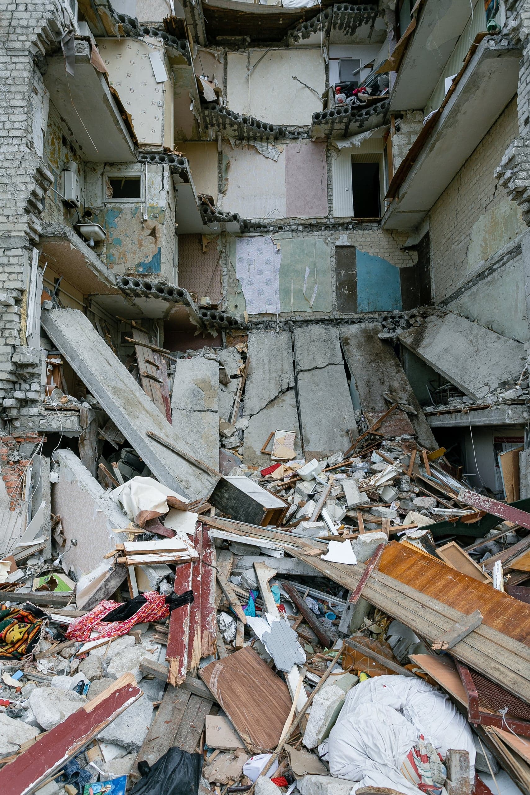Сильно разрушенный многоэтажный дом, обнаживший внутренние помещения с остатками обоев, и гора обломков, бетона и мусора у основания.