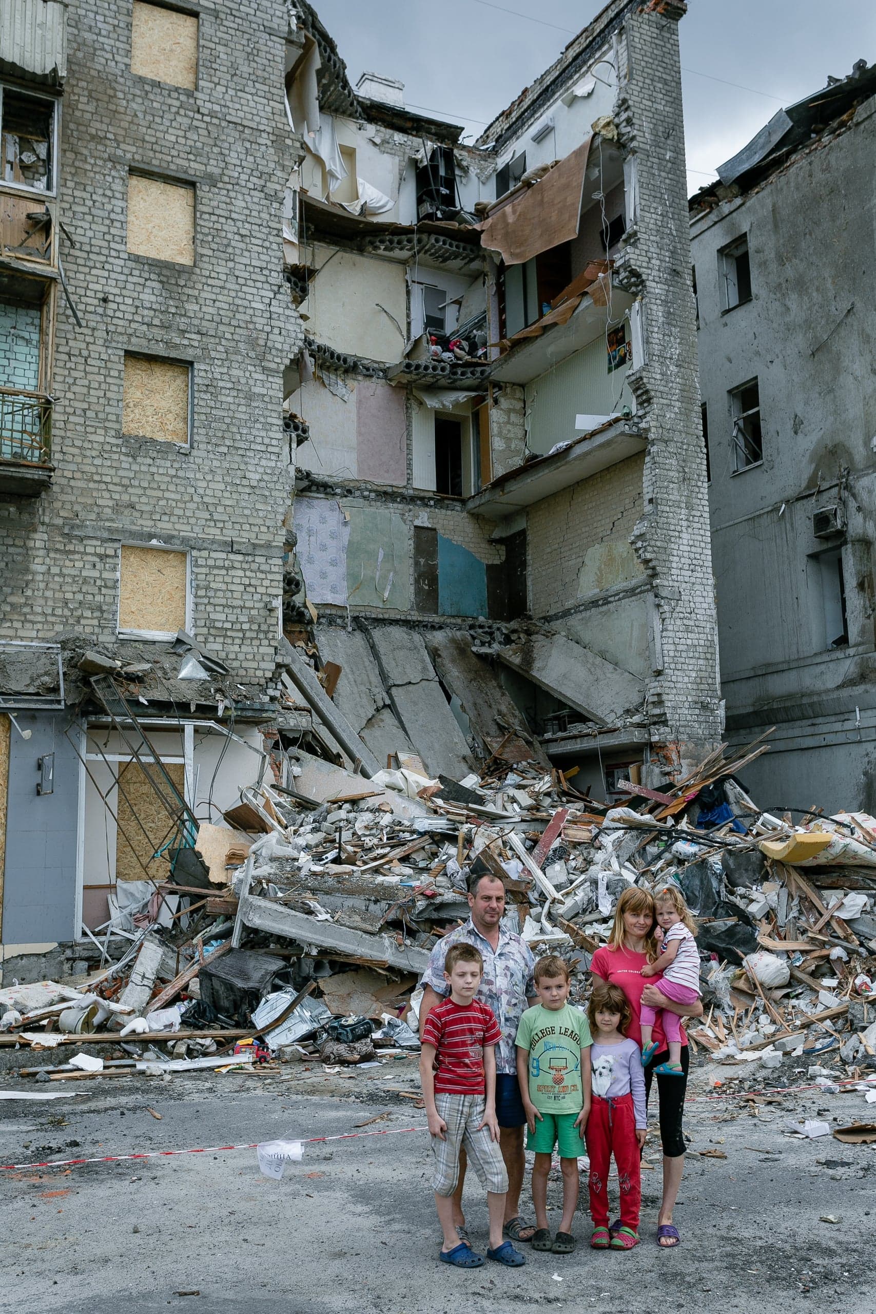 Семья из шести человек на фоне груды обломков и разрушенного многоэтажного дома.