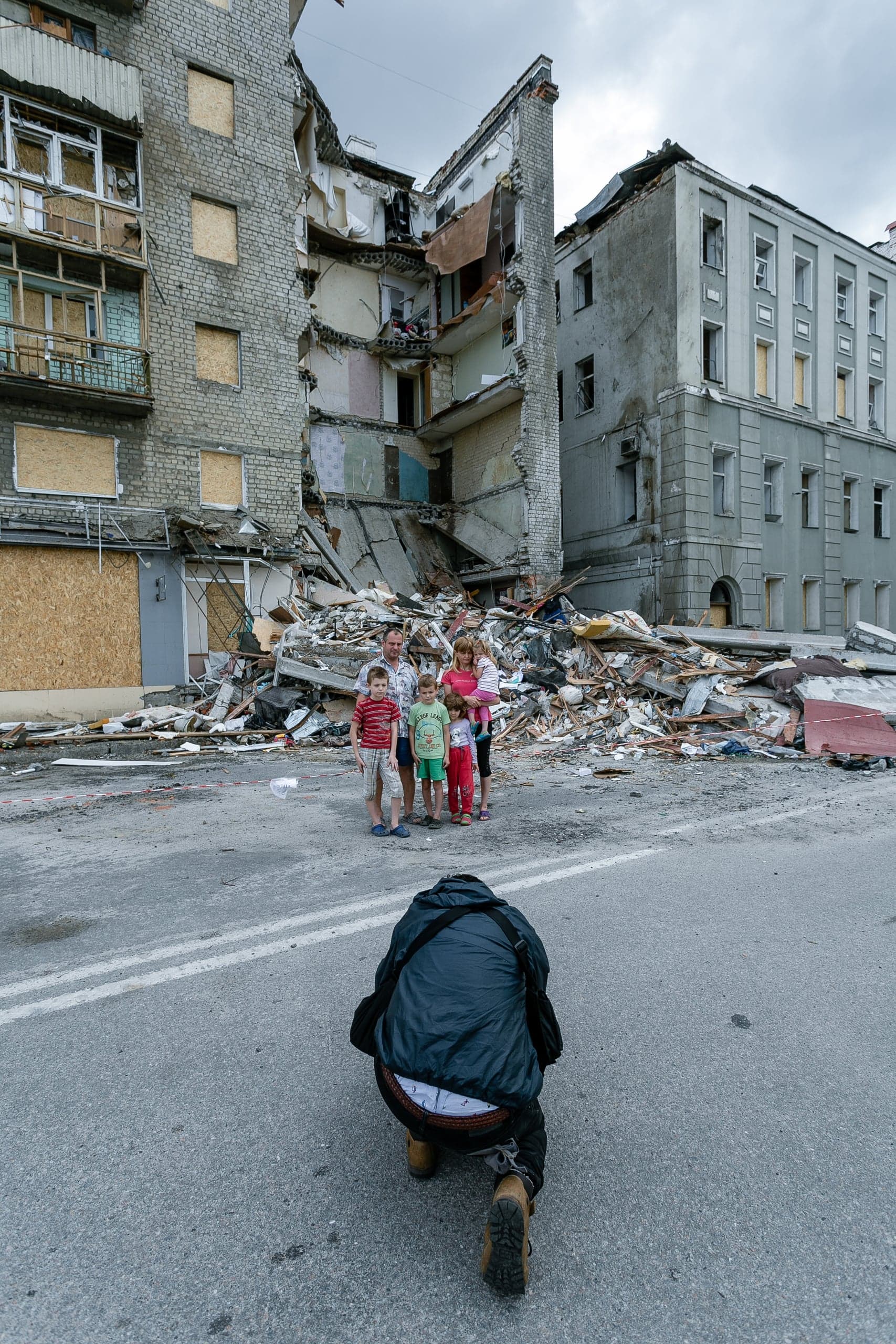 Фотограф снимает семью (двух взрослых и четырех детей) на фоне частично разрушенного взрывом многоэтажного жилого дома и руин.