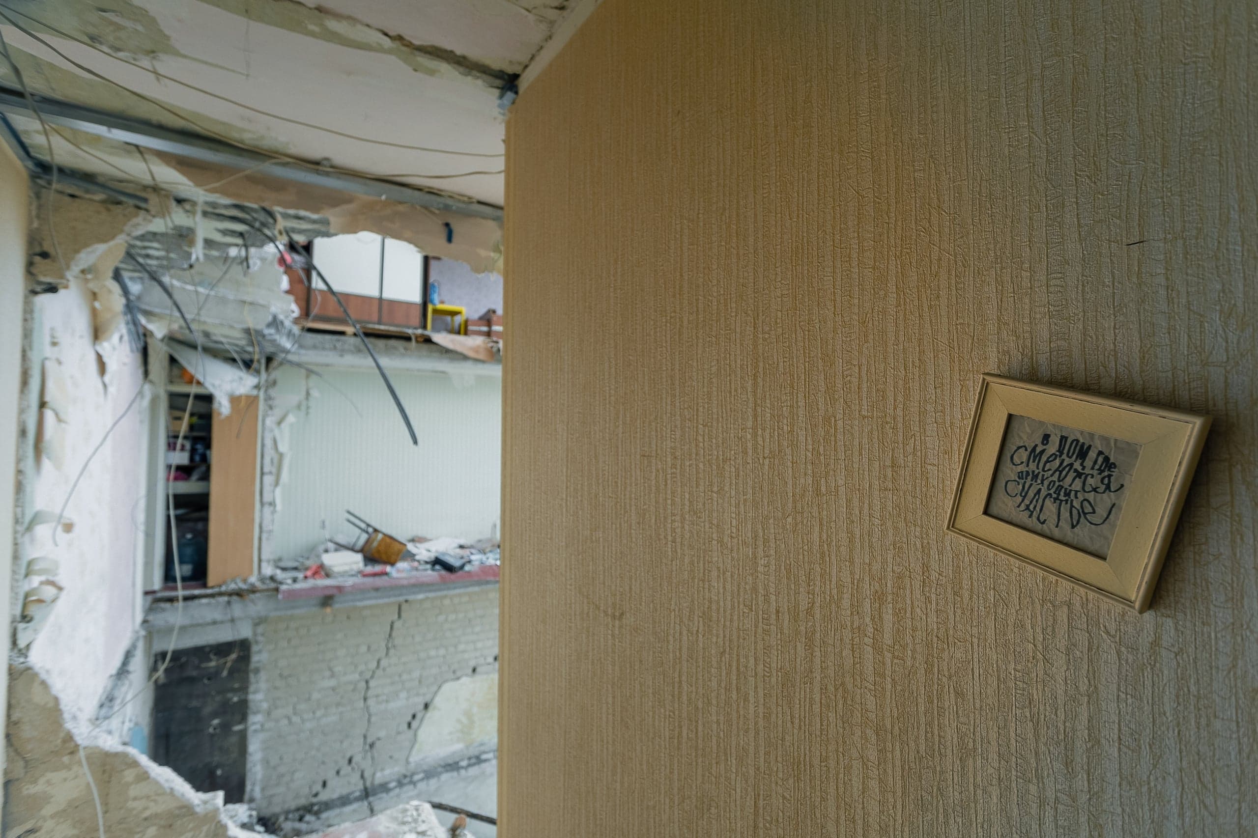 Поврежденная взрывом квартира, в глубине — обрушенные перекрытия и вид на разрушенное здание; справа на стене висит рамка с надписью: "В дом, где смеются, приходит счастье".