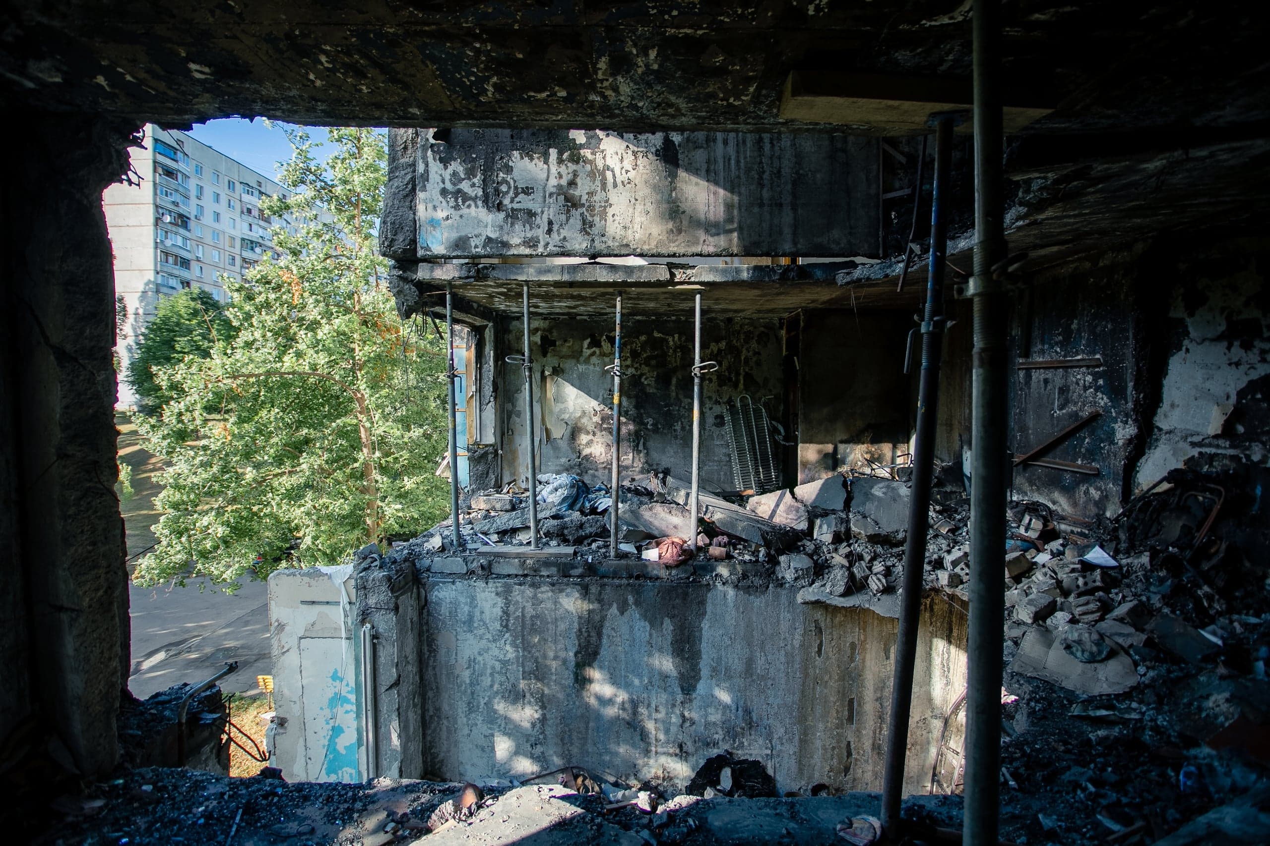 Разрушенная, выгоревшая квартира многоэтажного дома с обвалившимися перекрытиями и мусором; вид наружу на зелень и уцелевший жилой дом.