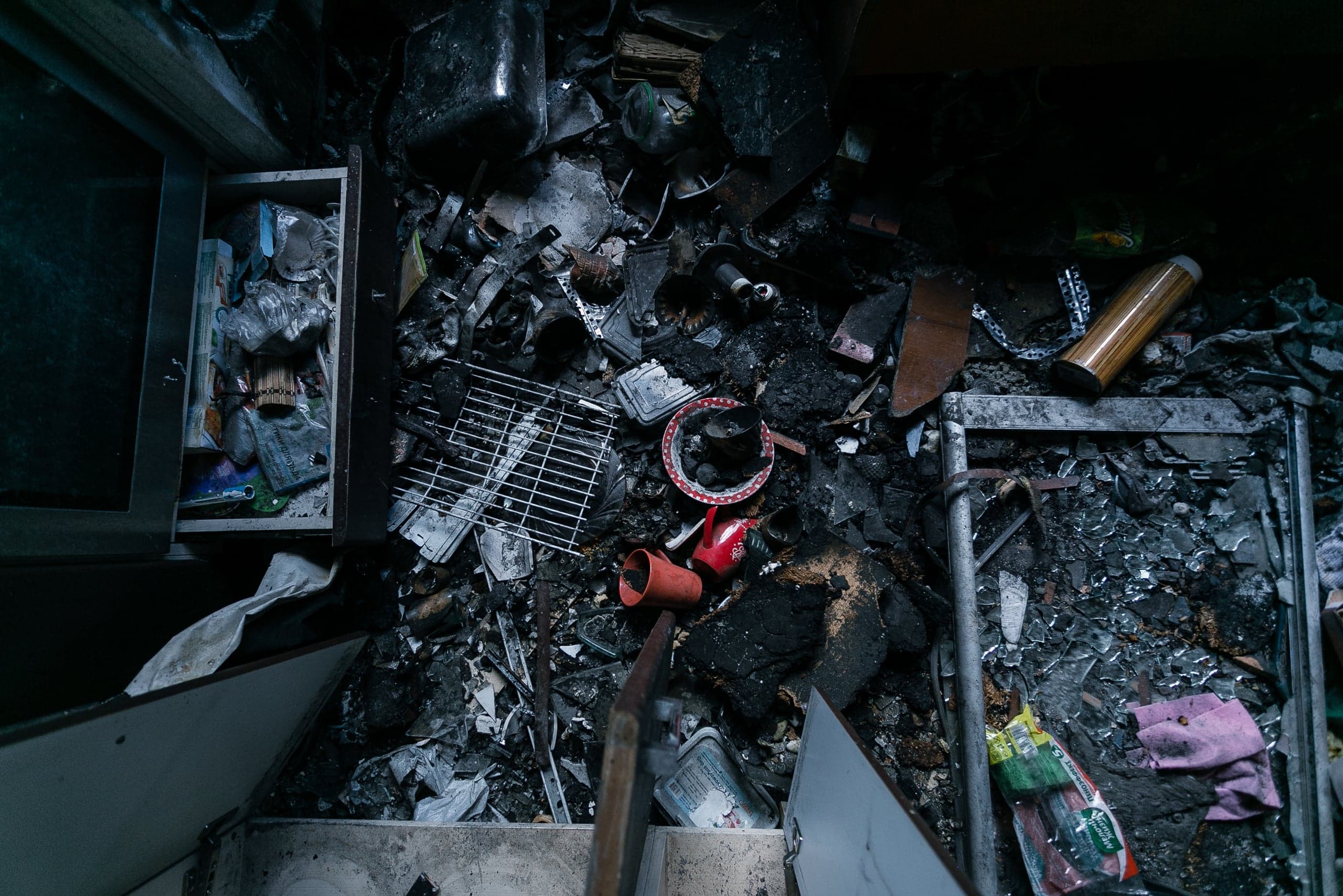 Последствия пожара: обугленный мусор, сломанная мебель, пепел и обломки в темном помещении.