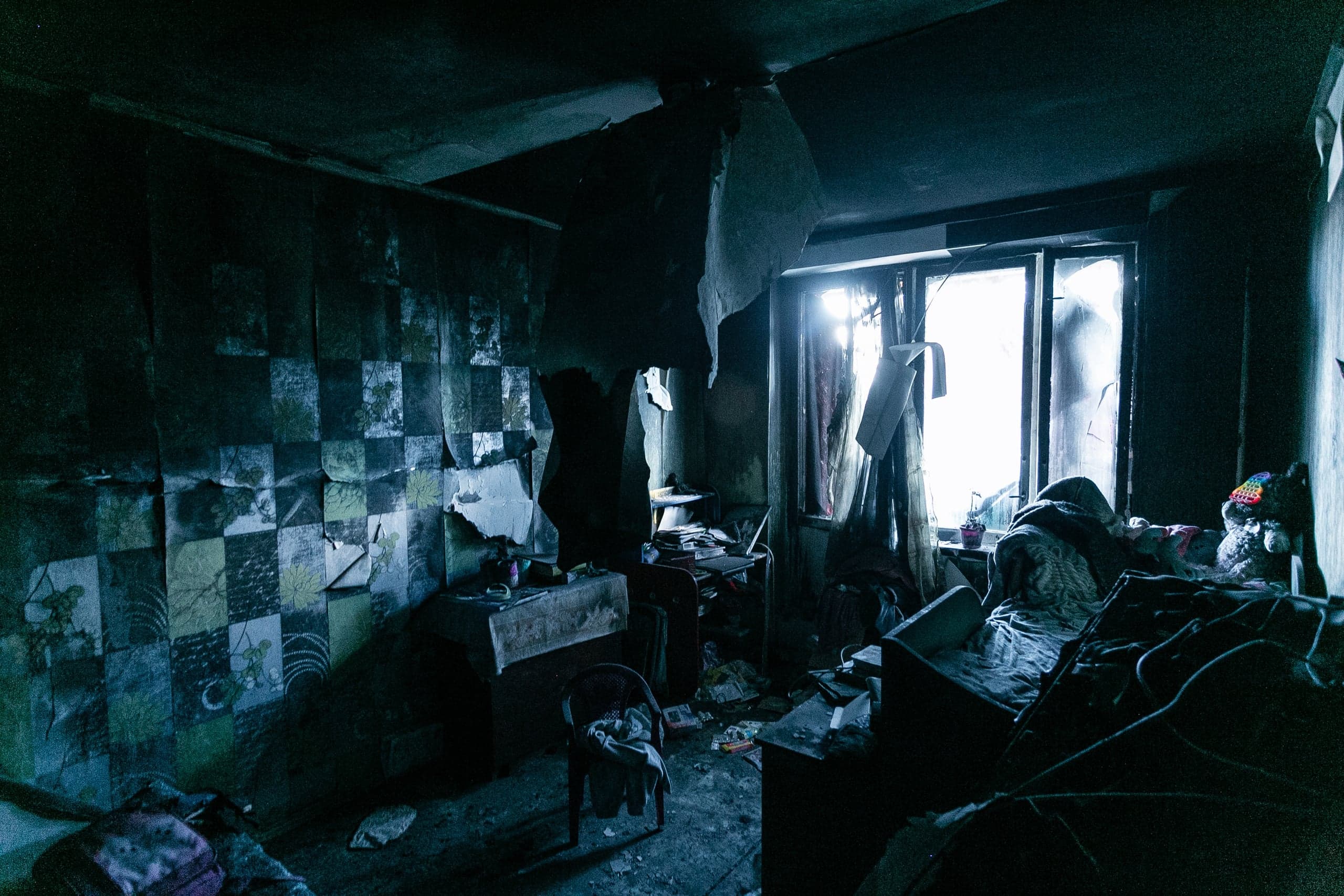 Разрушенная комната с обвалившимся потолком и оторванными обоями. Темный интерьер освещен ярким светом из окна; виден беспорядок и мягкая игрушка.