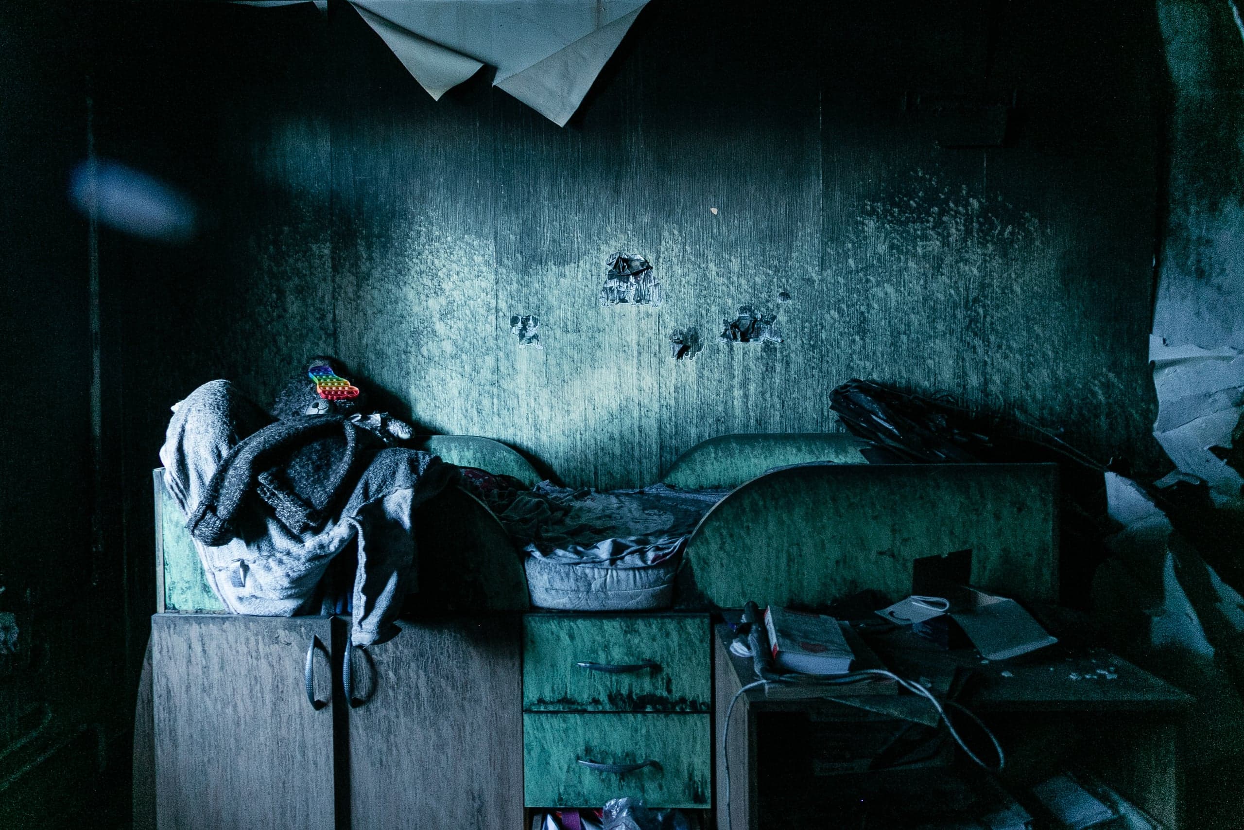 Затемненный интерьер заброшенной комнаты с облупившимися сине-зелеными стенами. В центре — низкий шкаф и диван, заваленные вещами.