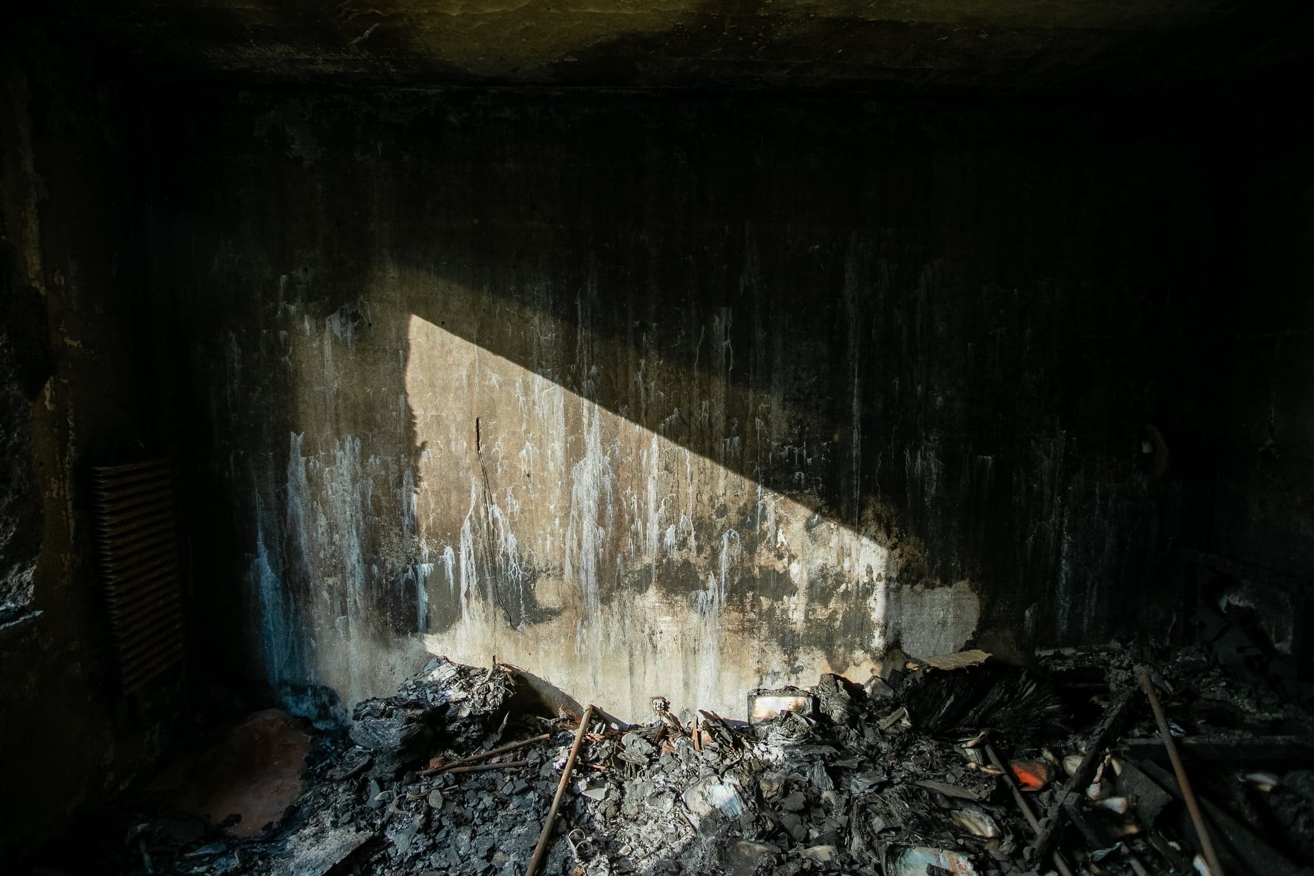 Внутренний угол разрушенного помещения с обугленными стенами, на которых видна белая грязь и треугольный луч света. Пол завален черным пеплом и обломками.
