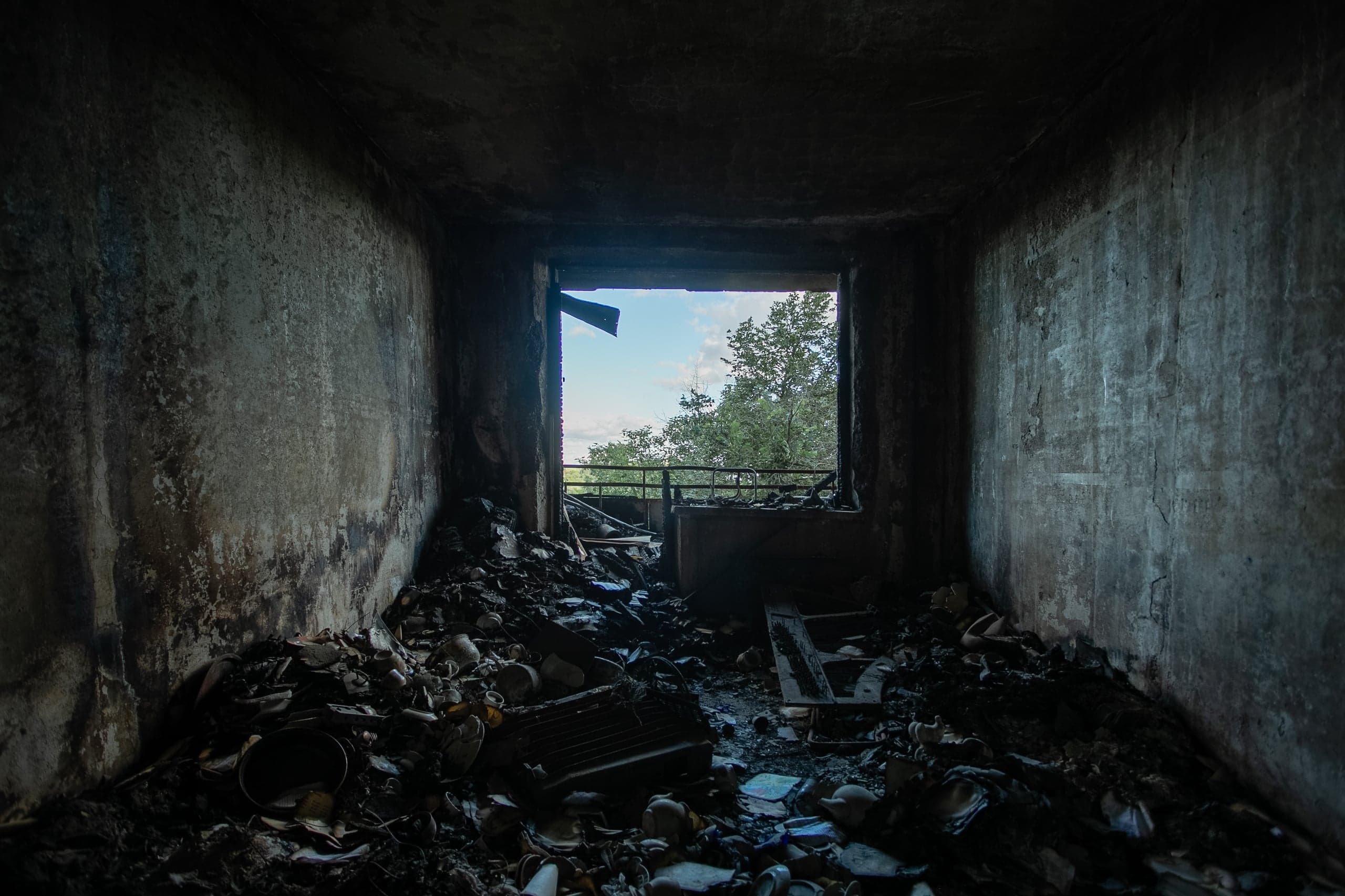 Сгоревшая, задымленная комната, заполненная мусором и обломками, с видом на зелёные деревья и синее небо через пустой дверной проём.