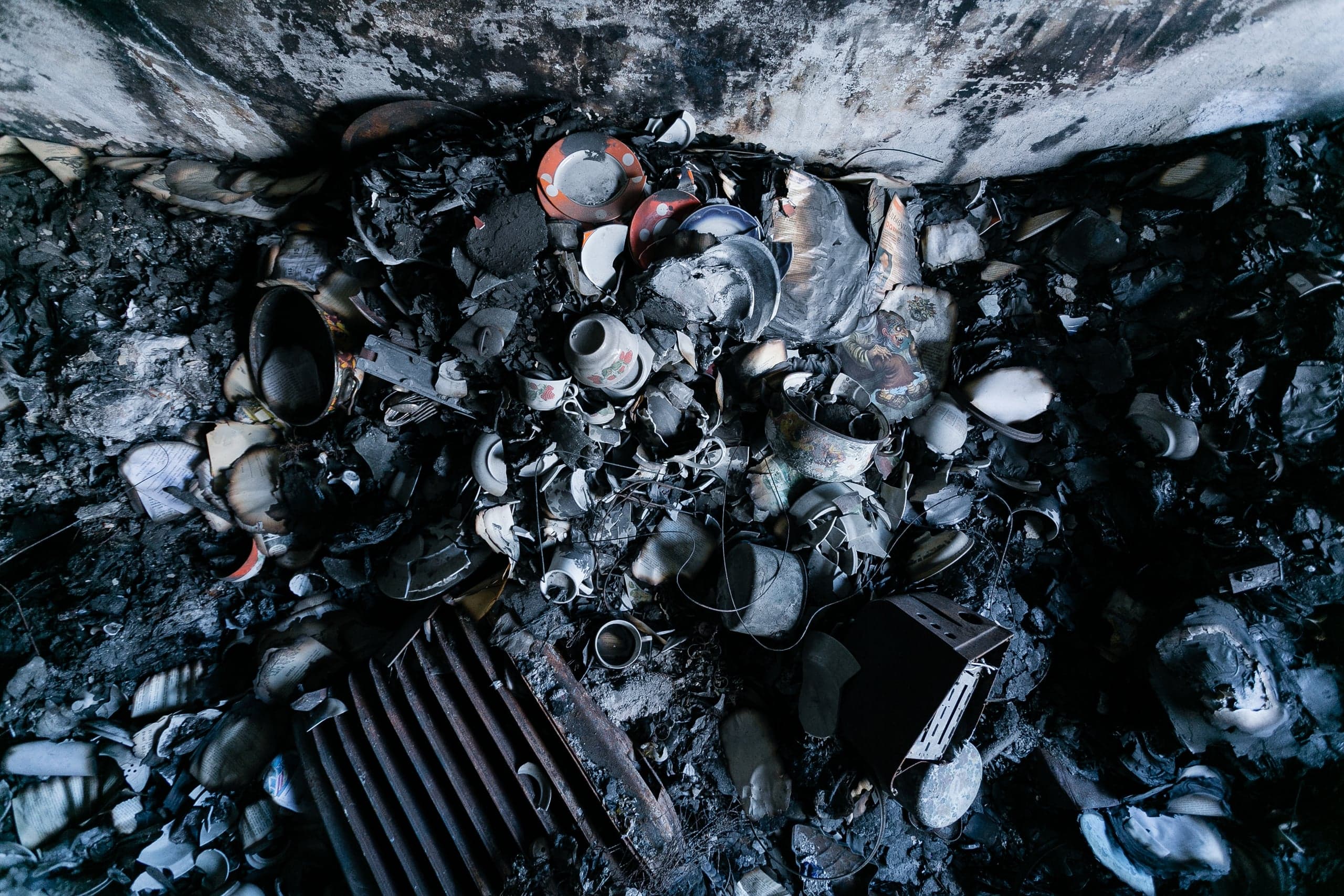 SOPA: Сгоревшая и разбитая домашняя утварь, посуда и обугленные предметы в углу разрушенного помещения.