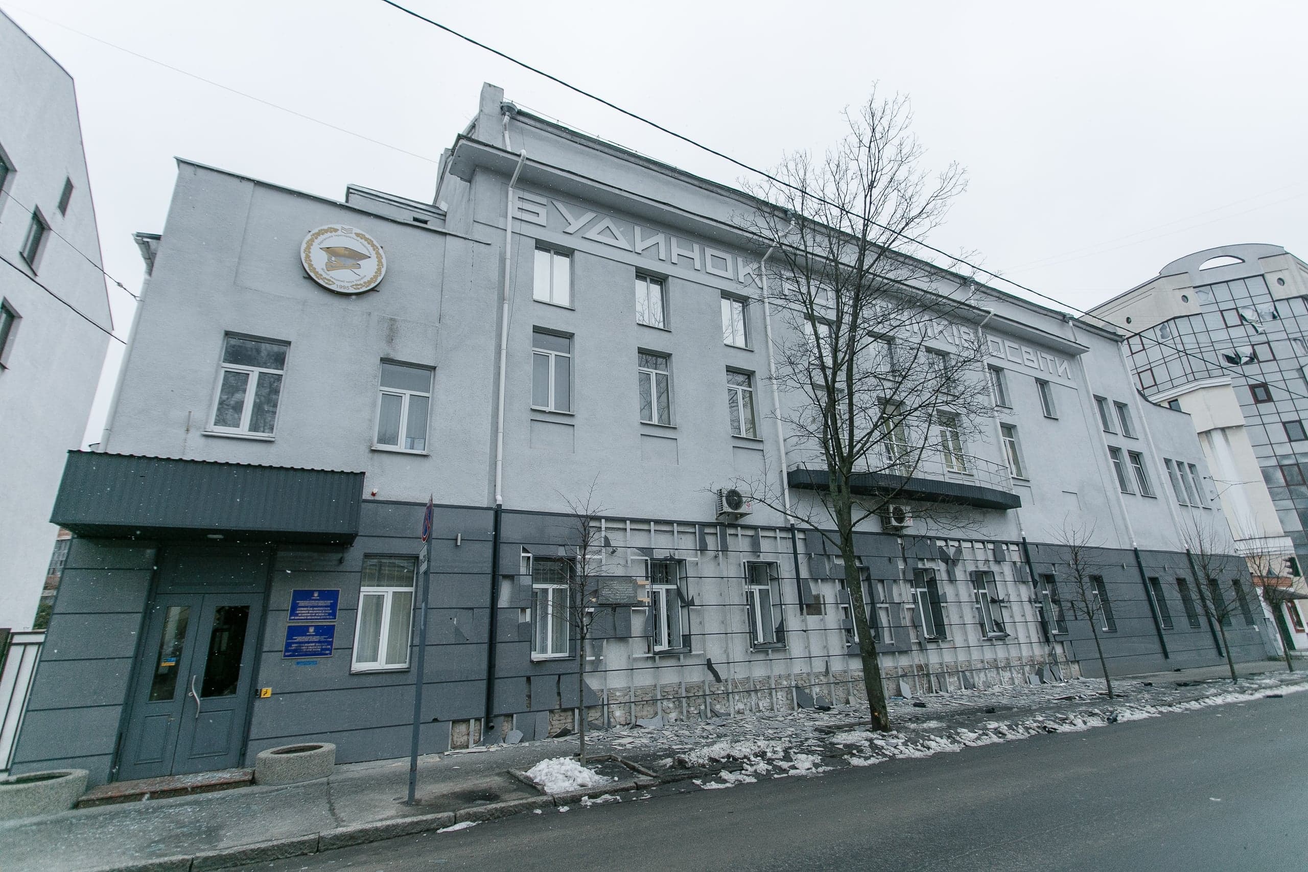 Серое здание с облупившимся фасадом нижнего этажа, над которым виден текст: "БУДИНОК ... ОСВІТИ". Зимний городской пейзаж.